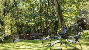 A serene outdoor seating area with a wooden bench and metal chairs, surrounded by lush greenery and trees.