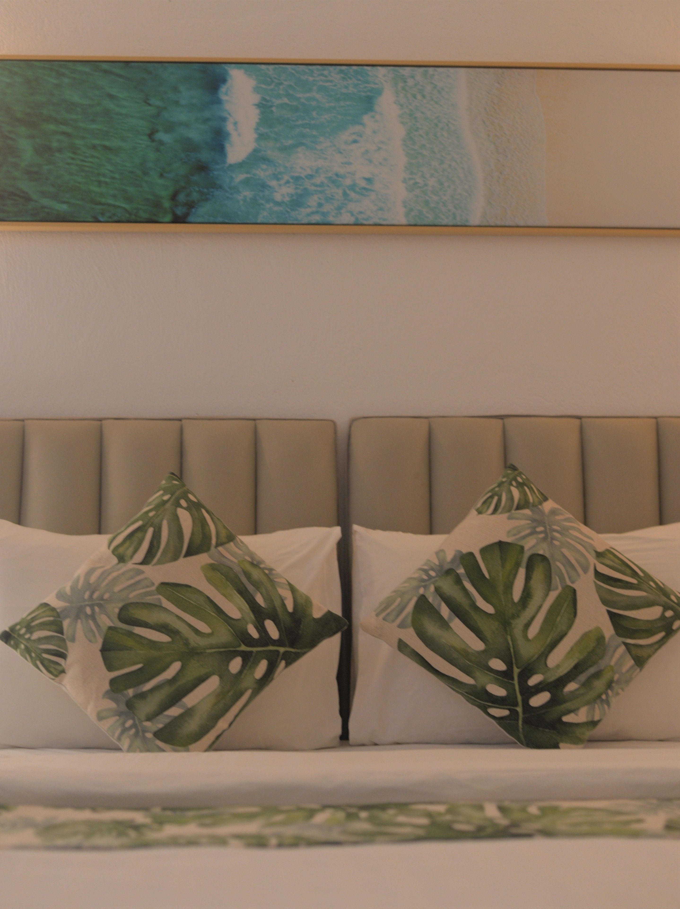 A bed with a headboard, two pillows with a leaf pattern, and a painting of a blue ocean above the bed.