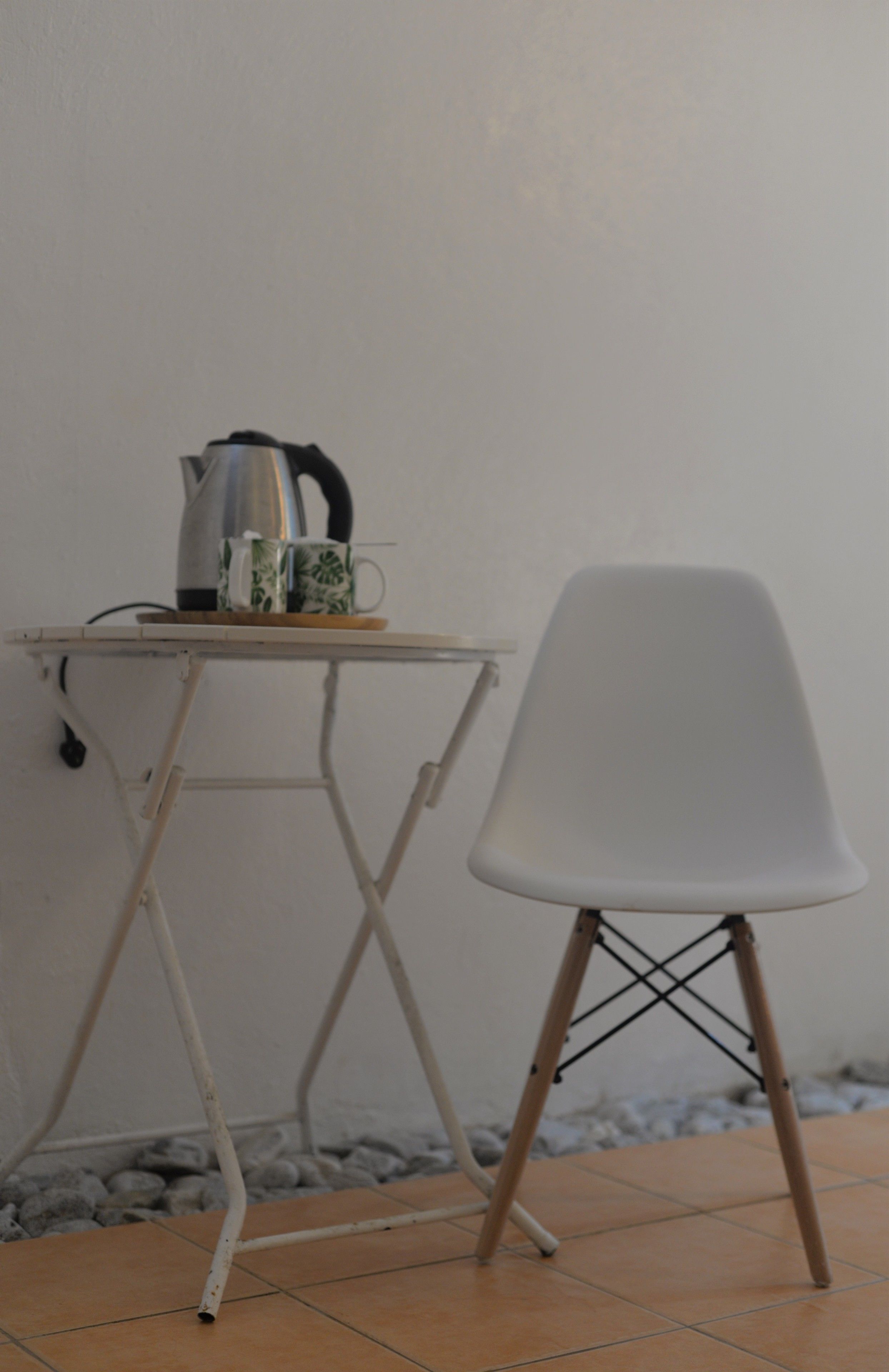 A small table with a kettle and a mug, a white chair, and a tiled floor.