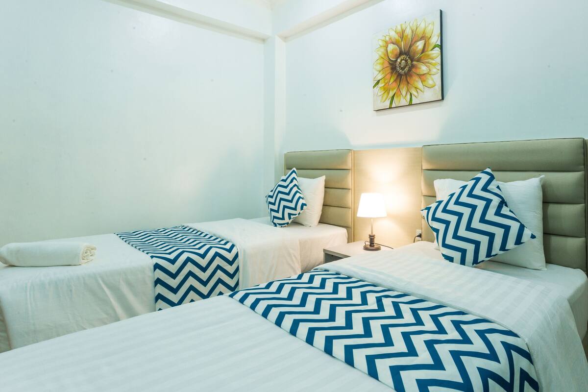 Hotelkamer met twee bedden, elk voorzien van een wit- en blauw gestreept dekbedovertrek, een beige hoofdbord en een wandlamp.