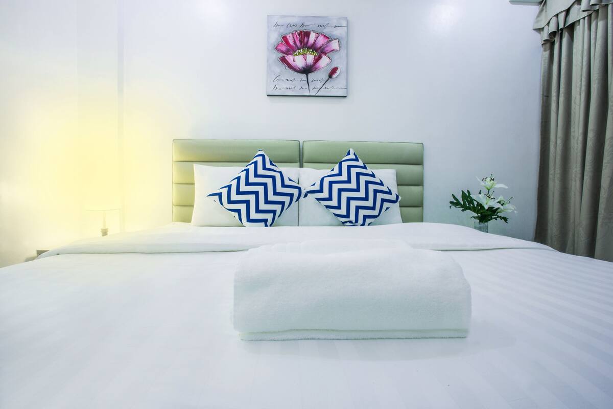 Een opgemaakt bed met een groen hoofdeinde, twee kussens met een blauw zigzagpatroon, en een witte handdoek gevouwen aan het voeteneinde.