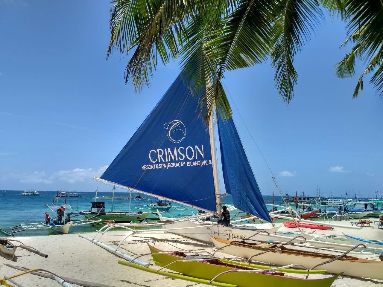 A beach with a sailboat named 'CRIMSON' docked, surrounded by smaller boats and palm trees.