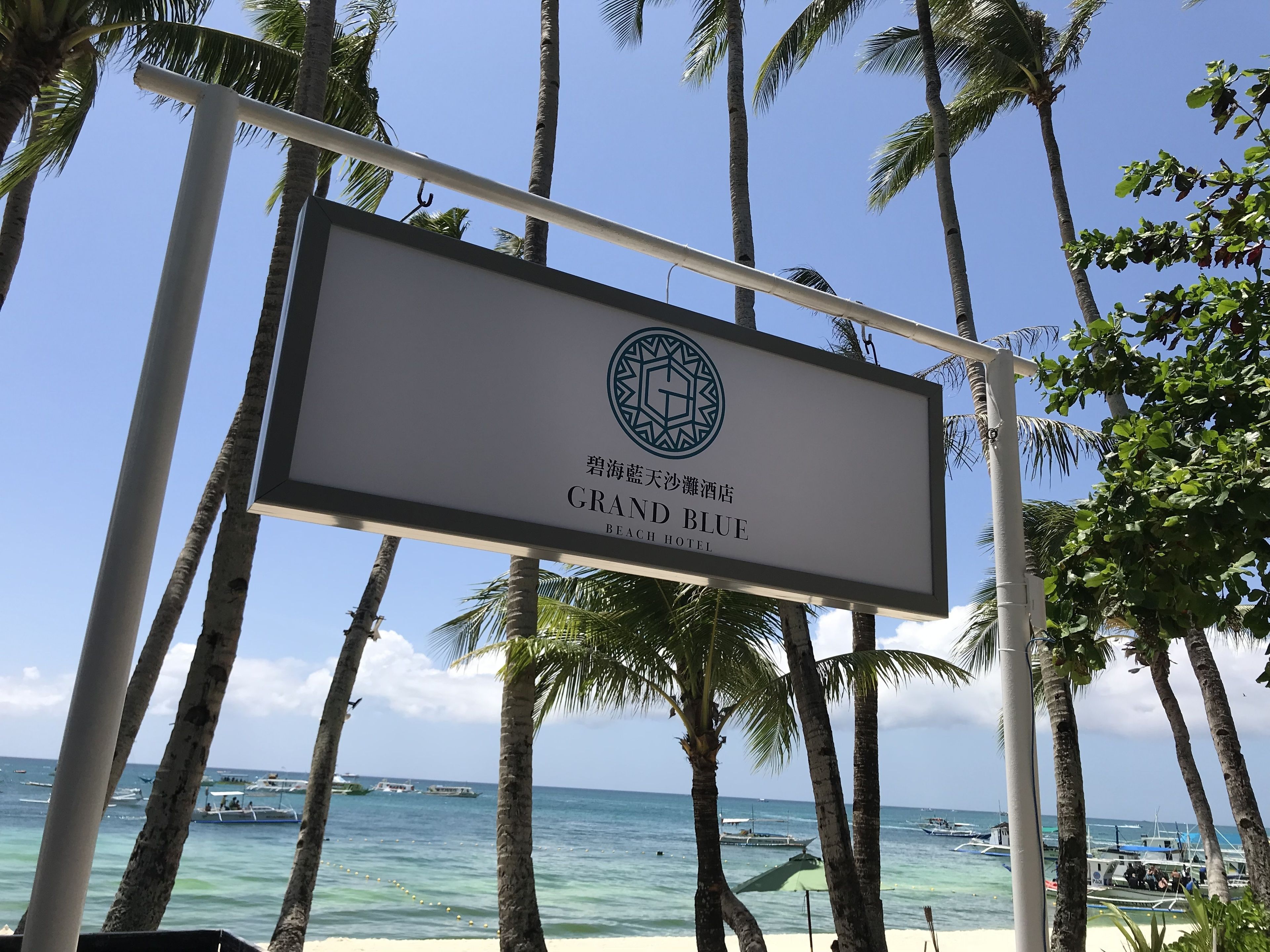 A sign for Grand Blue Beach Hotel with palm trees and boats in the background.