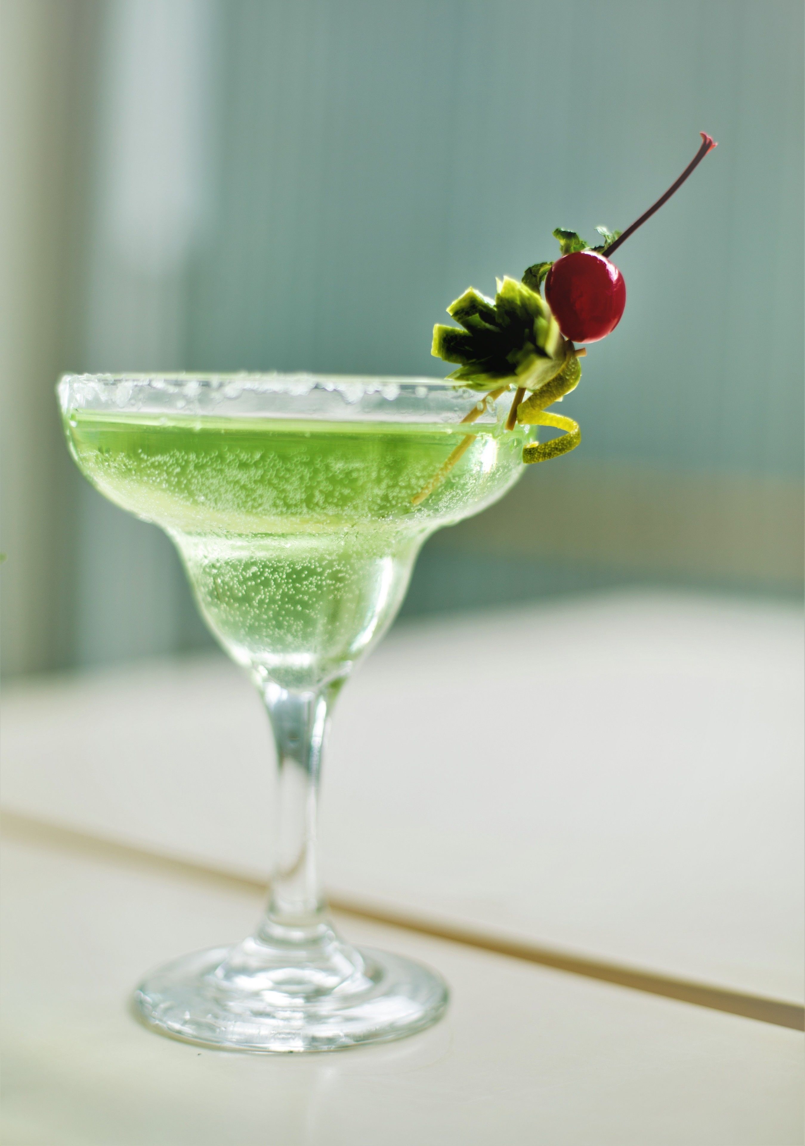 A martini glass with a green liquid, a cherry garnish, and a lemon twist on a white surface.