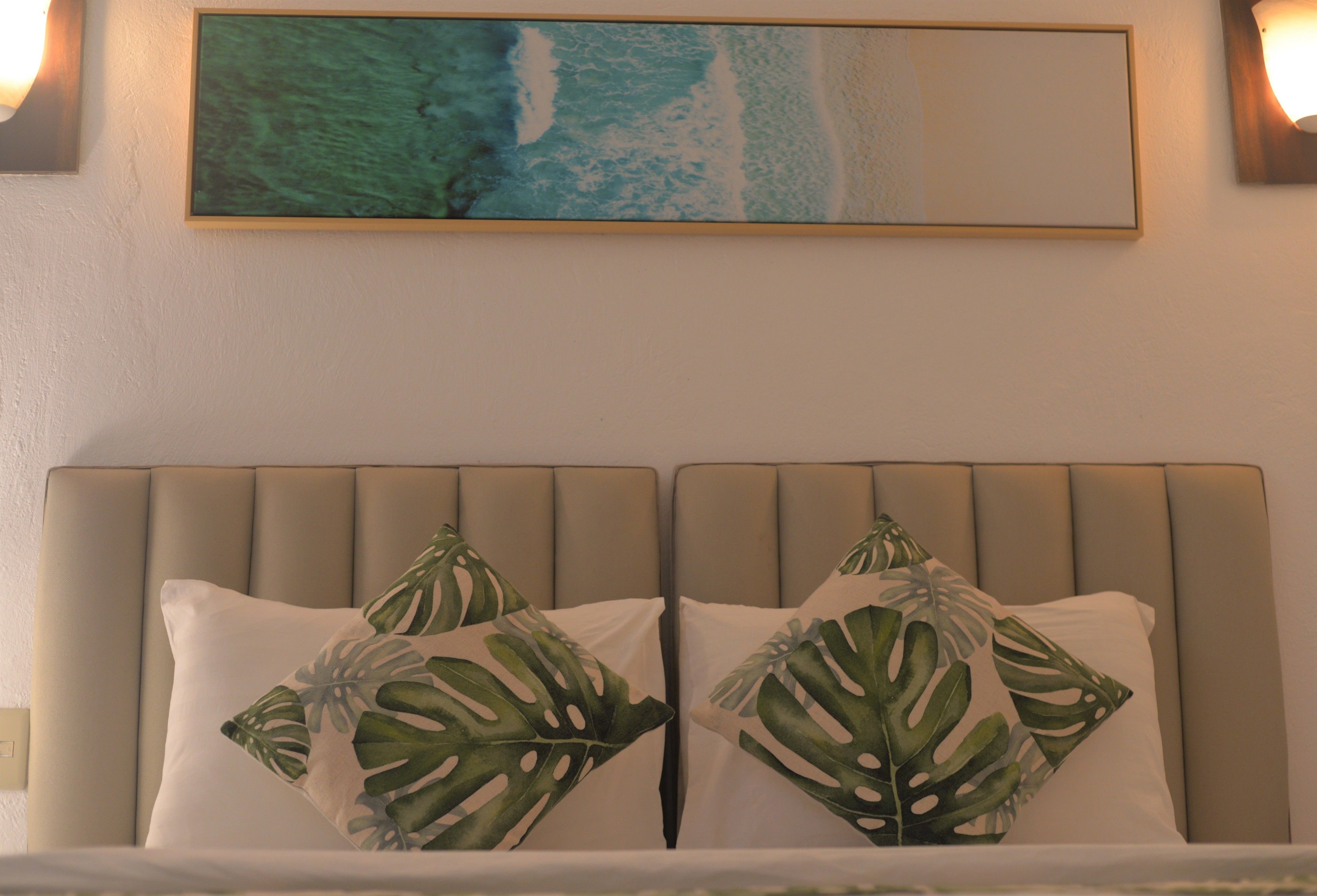 Una cama con cabecera, dos almohadas con estampado de hojas y un cuadro de una playa.