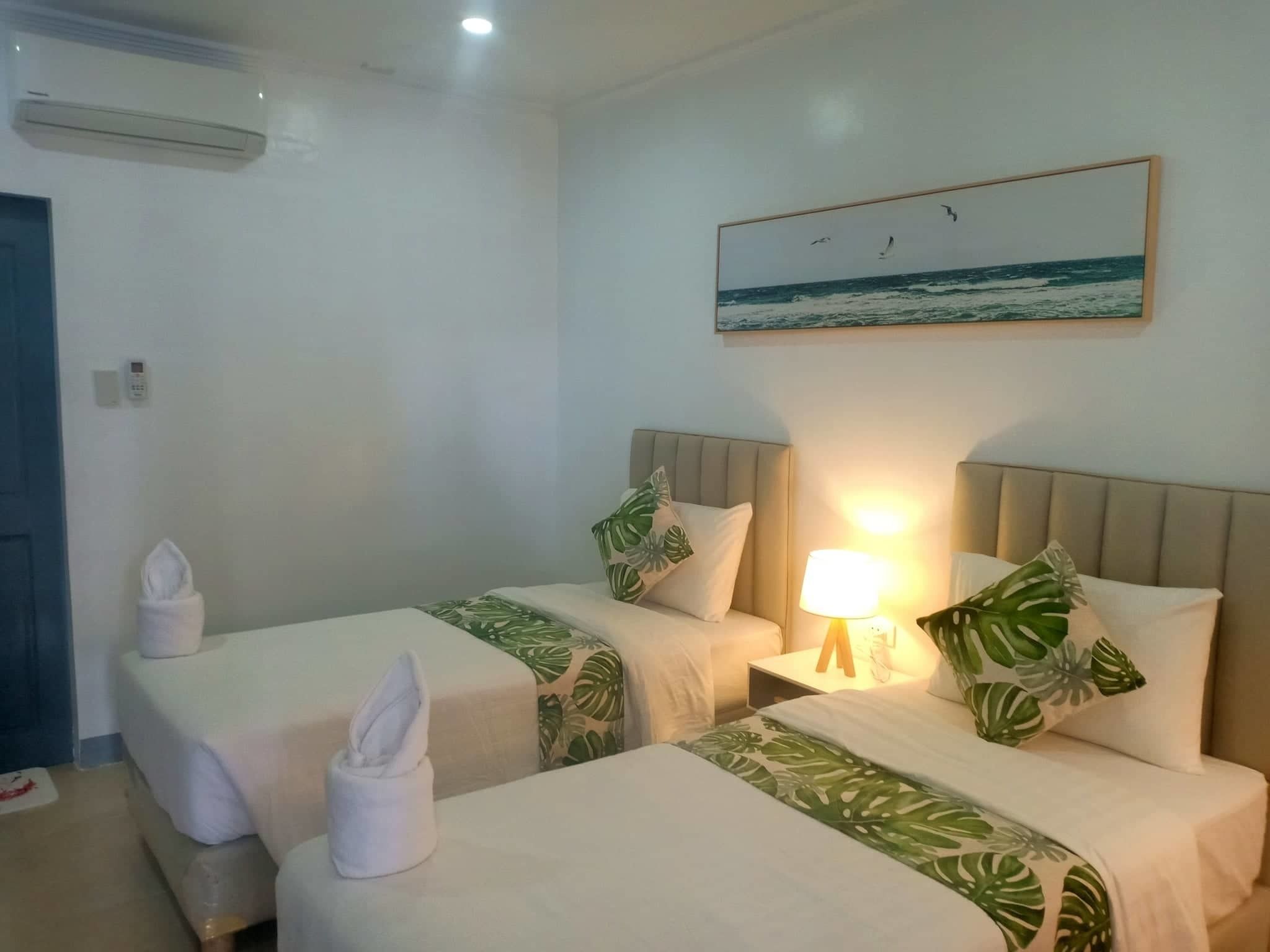 A hotel room with two beds, each with a green leaf-patterned bedspread, a bedside lamp, and a painting of the ocean on the wall.