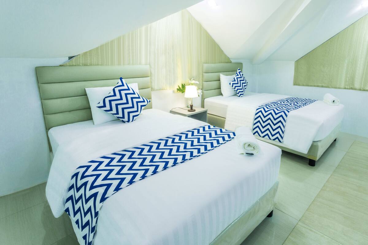 Een hotelkamer met twee bedden, elk met een witte sprei en blauwe zigzagkussens. Daartussen staat een nachtkastje met een lamp.