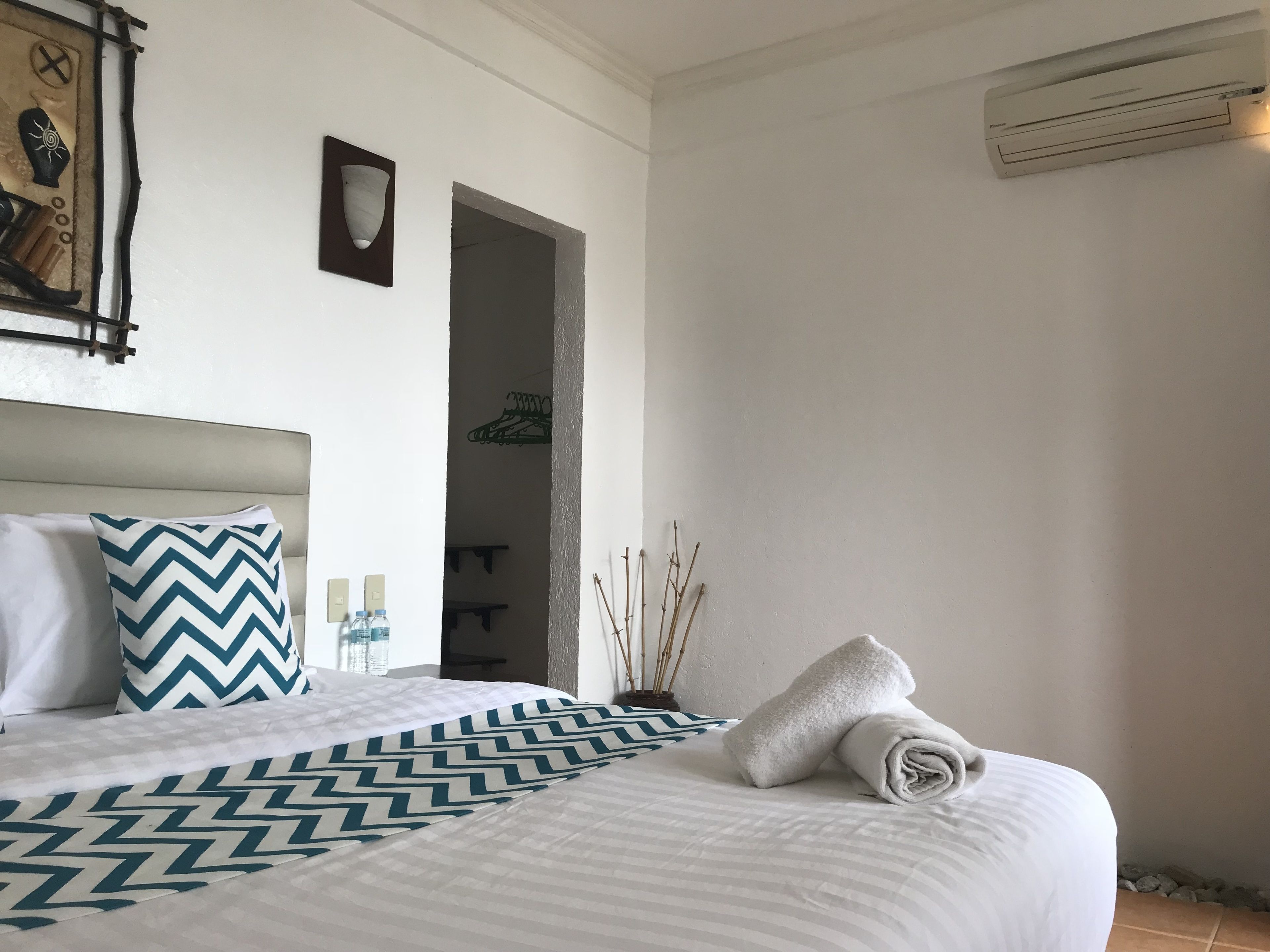 Een net opgemaakt bed met een beddekleed met patroon, een kussen en een opgerolde handdoek. Er is een wandairconditioner en een klein plankje met flessen.