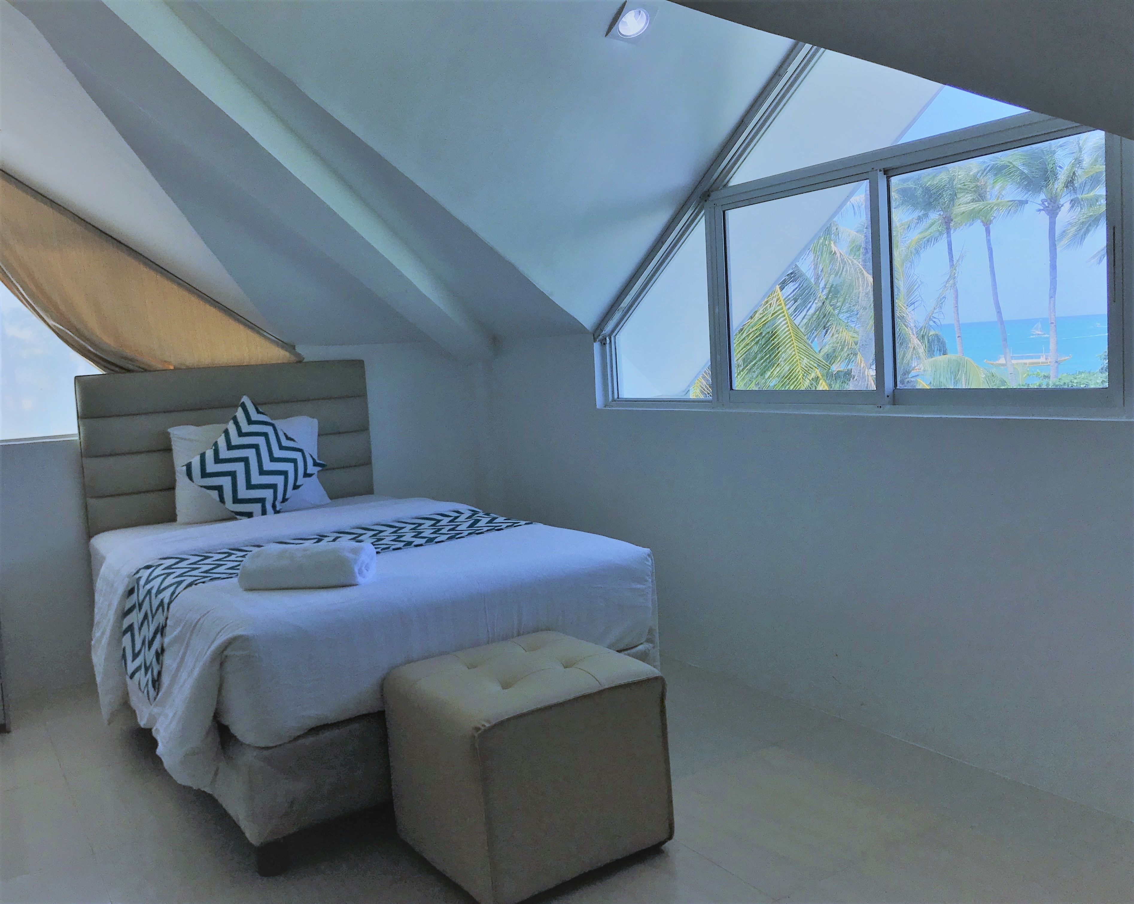 A modern bedroom with a large bed, a headboard, a window with a view of palm trees, and a skylight.