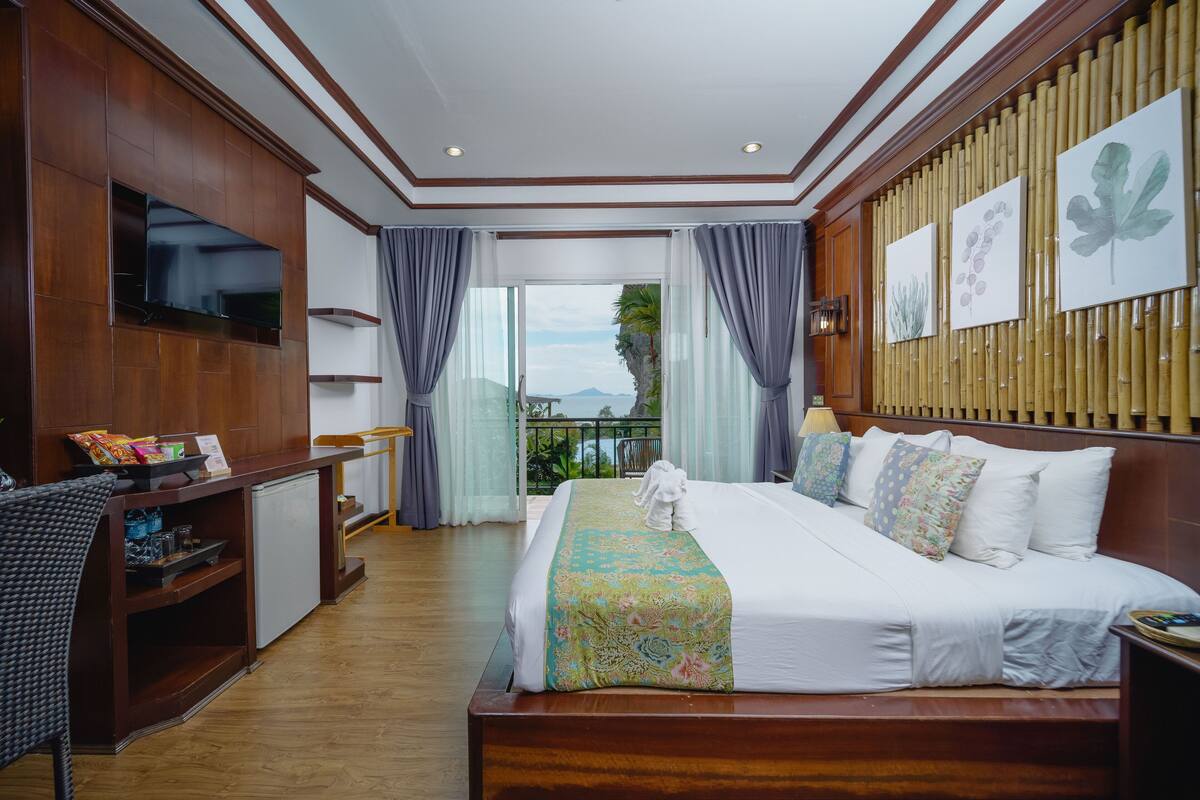 A hotel room with a large bed, a TV mounted on the wall, a desk with a computer, and a balcony with a view of the mountains.