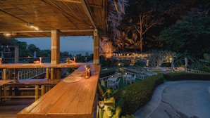 An outdoor dining area with wooden tables and chairs, surrounded by lush greenery and a view of a distant building. - Railay Phutawan Resort (Krabi)