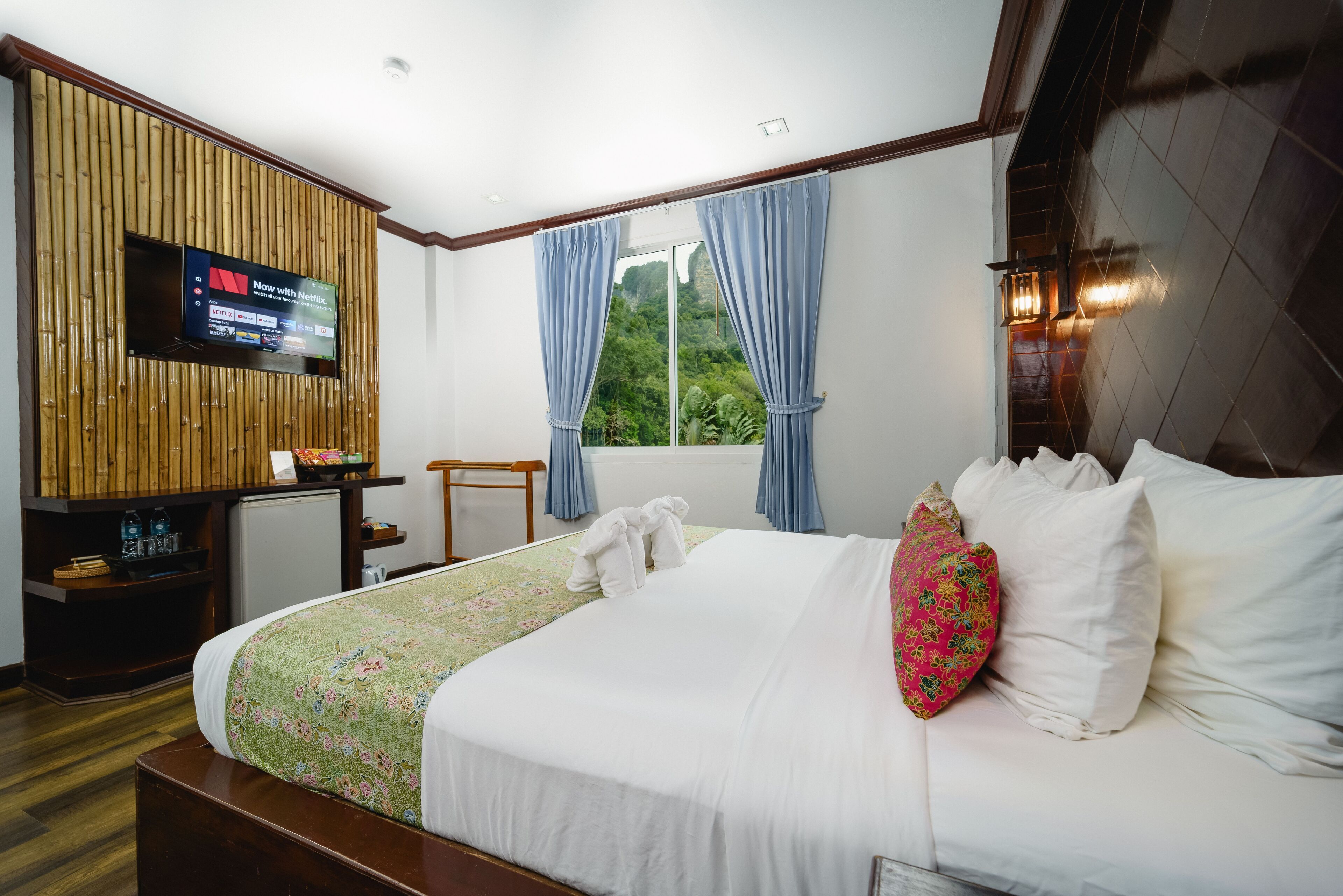 Habitación de hotel con cama, televisión montada en un panel de madera, una mesita y una ventana con cortinas.