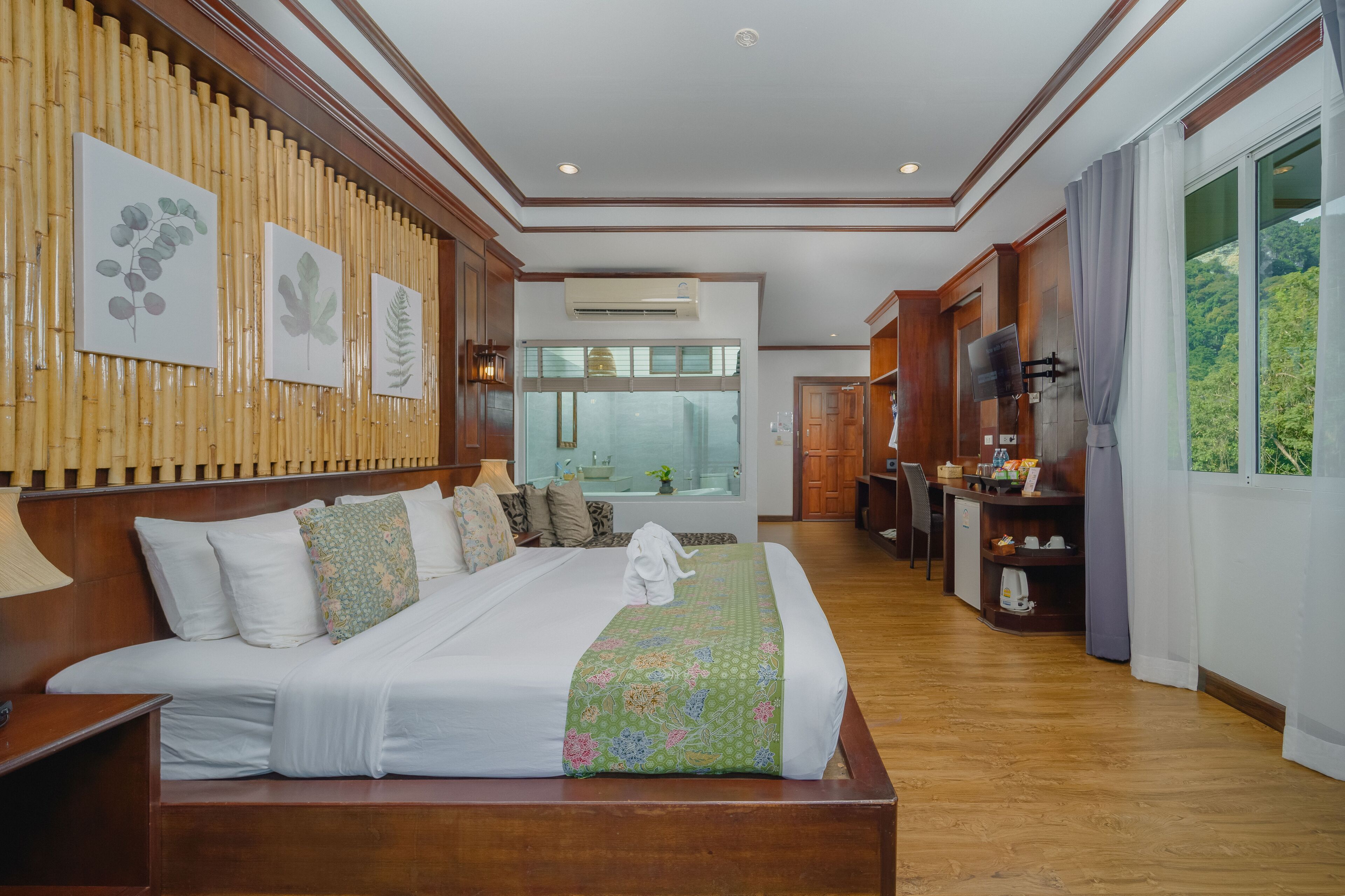 A hotel room with a large bed, a wooden headboard, a television, a desk with a chair, and a window with a view of greenery.