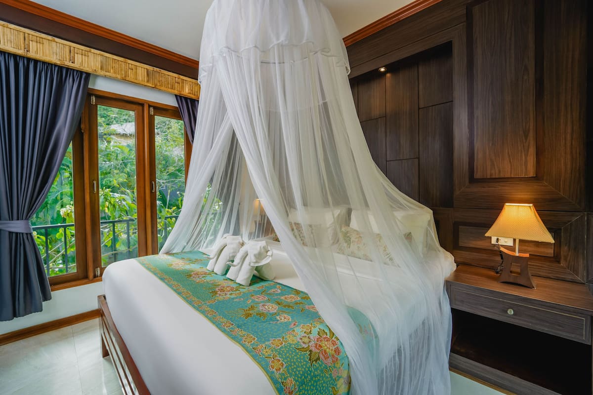 A bedroom with a bed under a canopy, a nightstand with a lamp, and a view of greenery through the window.
