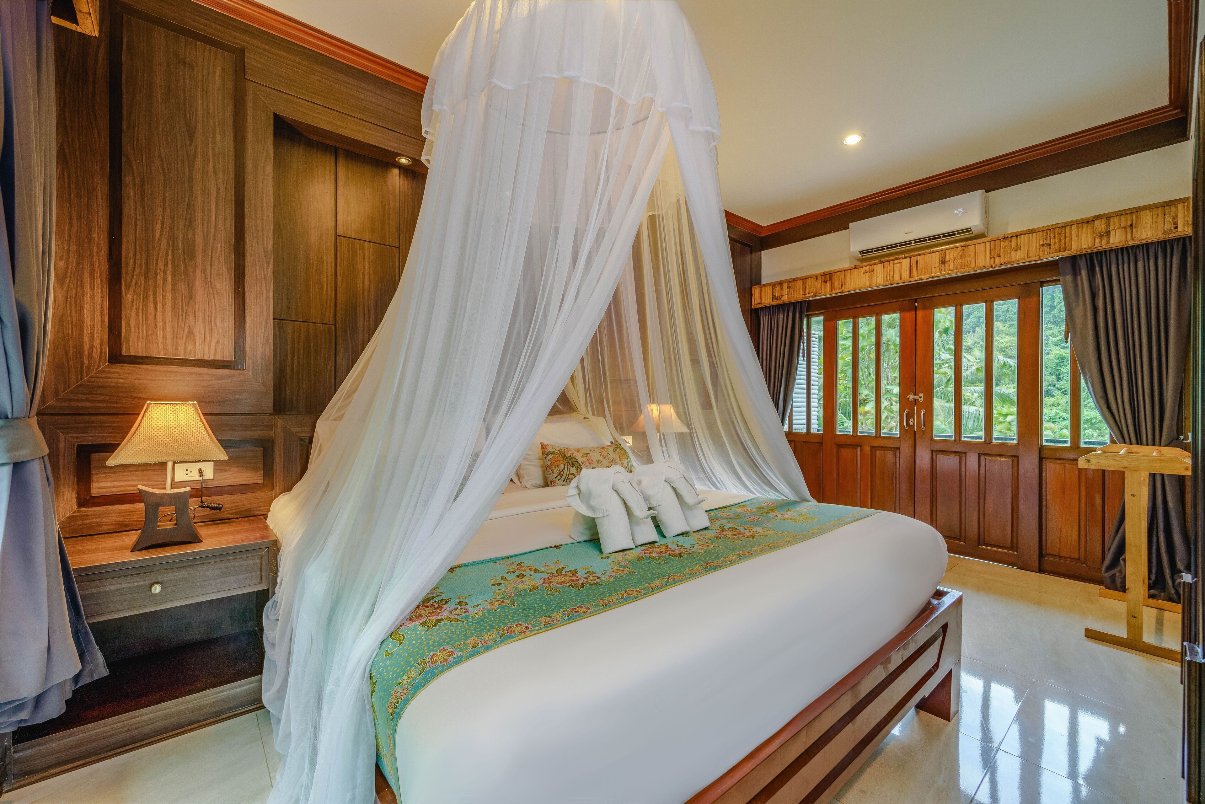 A bedroom with a canopy bed, wooden paneling, a bedside table with a lamp, and a view of greenery through large windows.