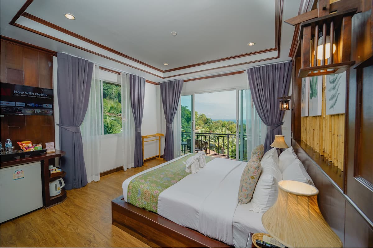 A hotel room with a large bed, a desk with a television, a balcony with a view, and a wooden wall.