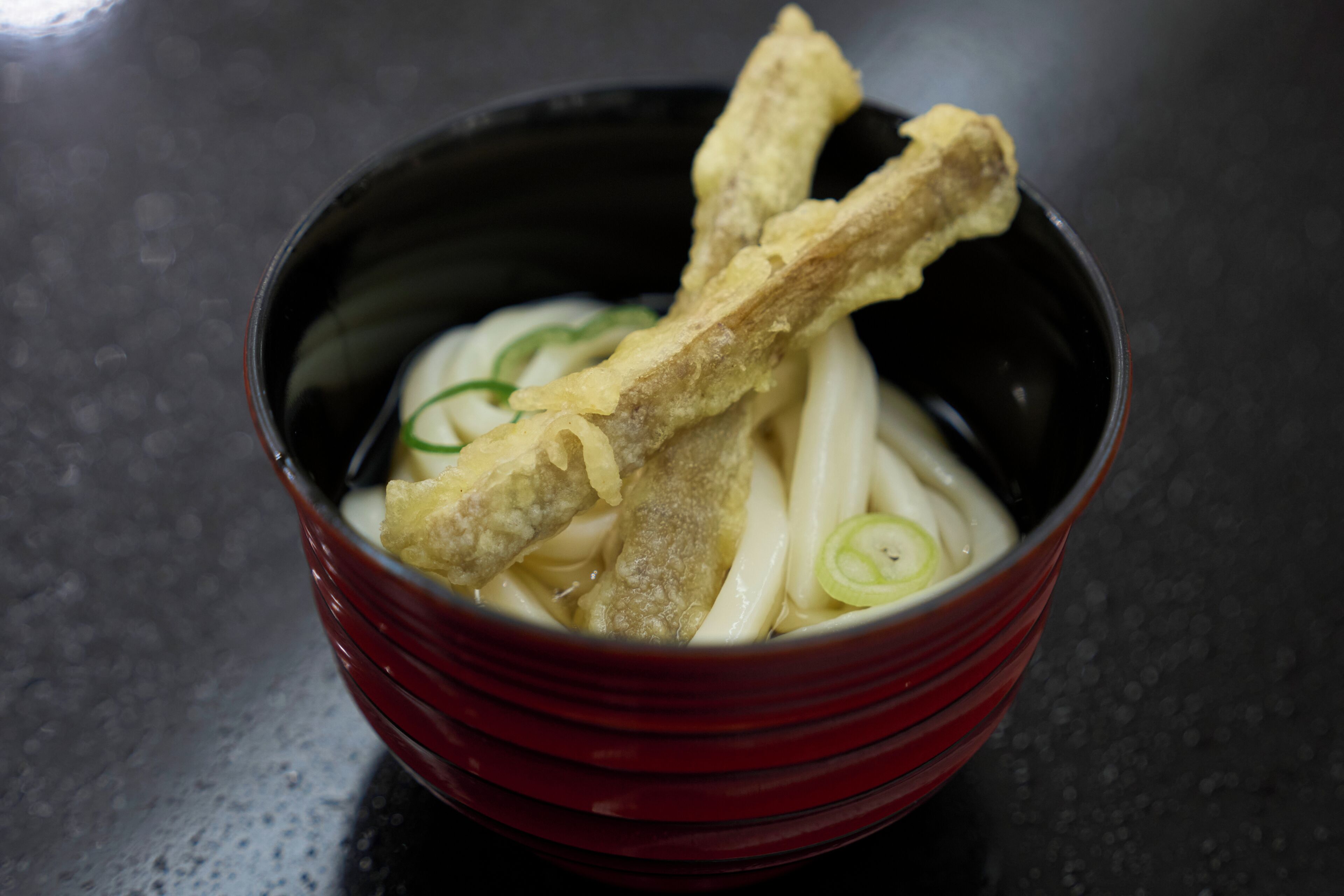 A bowl of noodles with fried items and green garnishes.