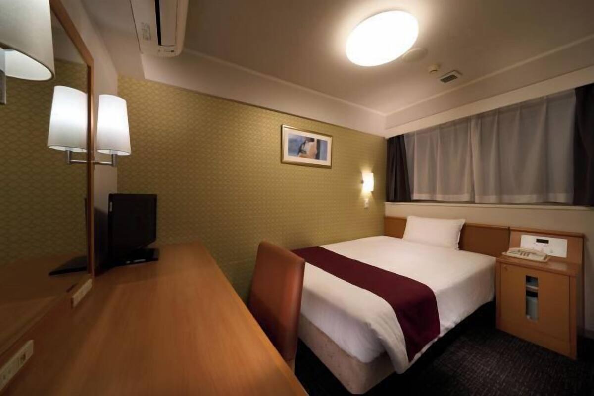 Hotellihuone, jossa on sänky, työpöytä, tuoli, televisio, yöpöytä, lamppu, taulu seinällä ja ikkuna, jossa on verhot.