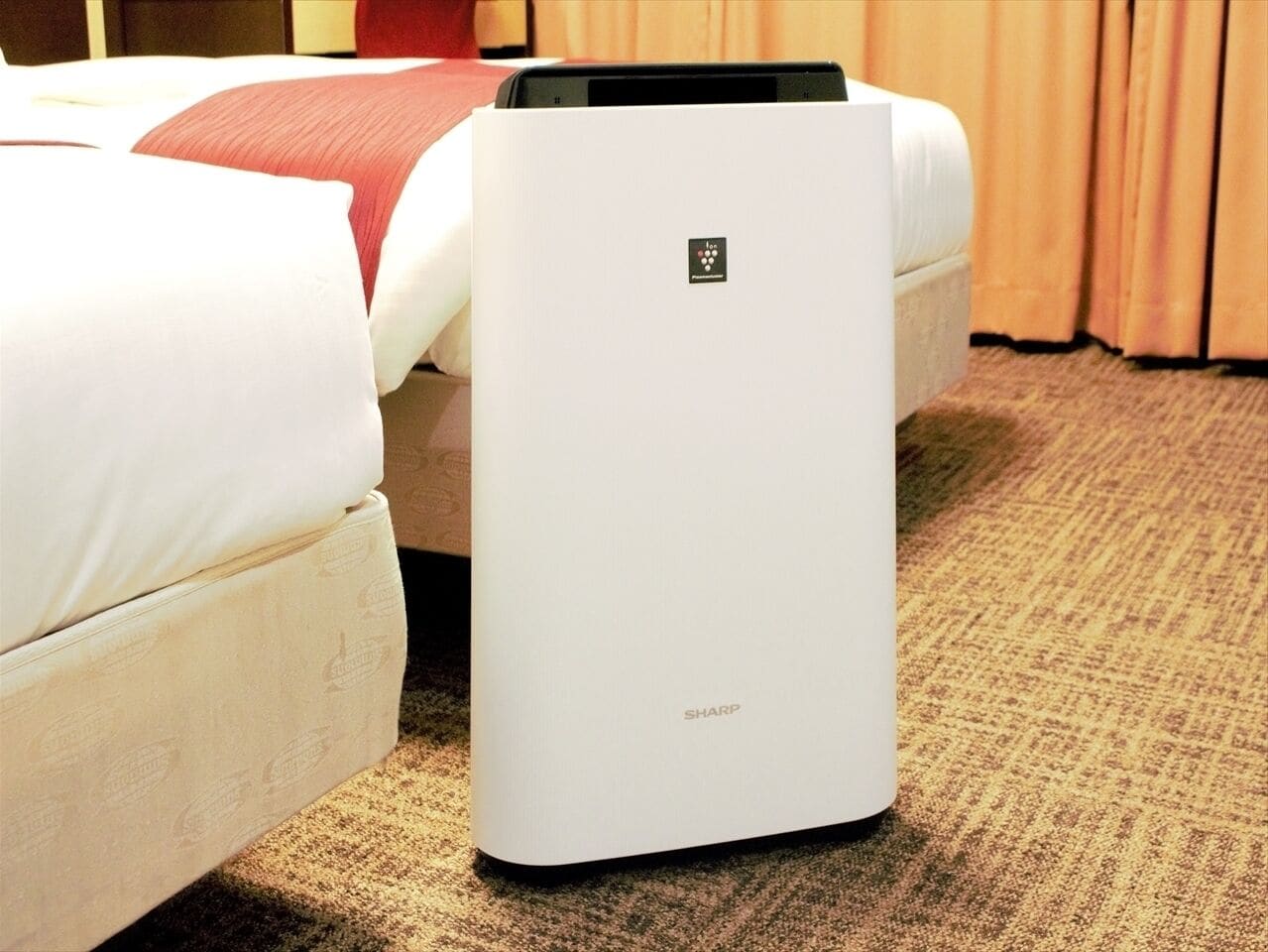 Ein weißes Sharp Luftreinigergerät in einem Hotelzimmer mit Teppichboden und einem Bett mit weißer Bettwäsche.