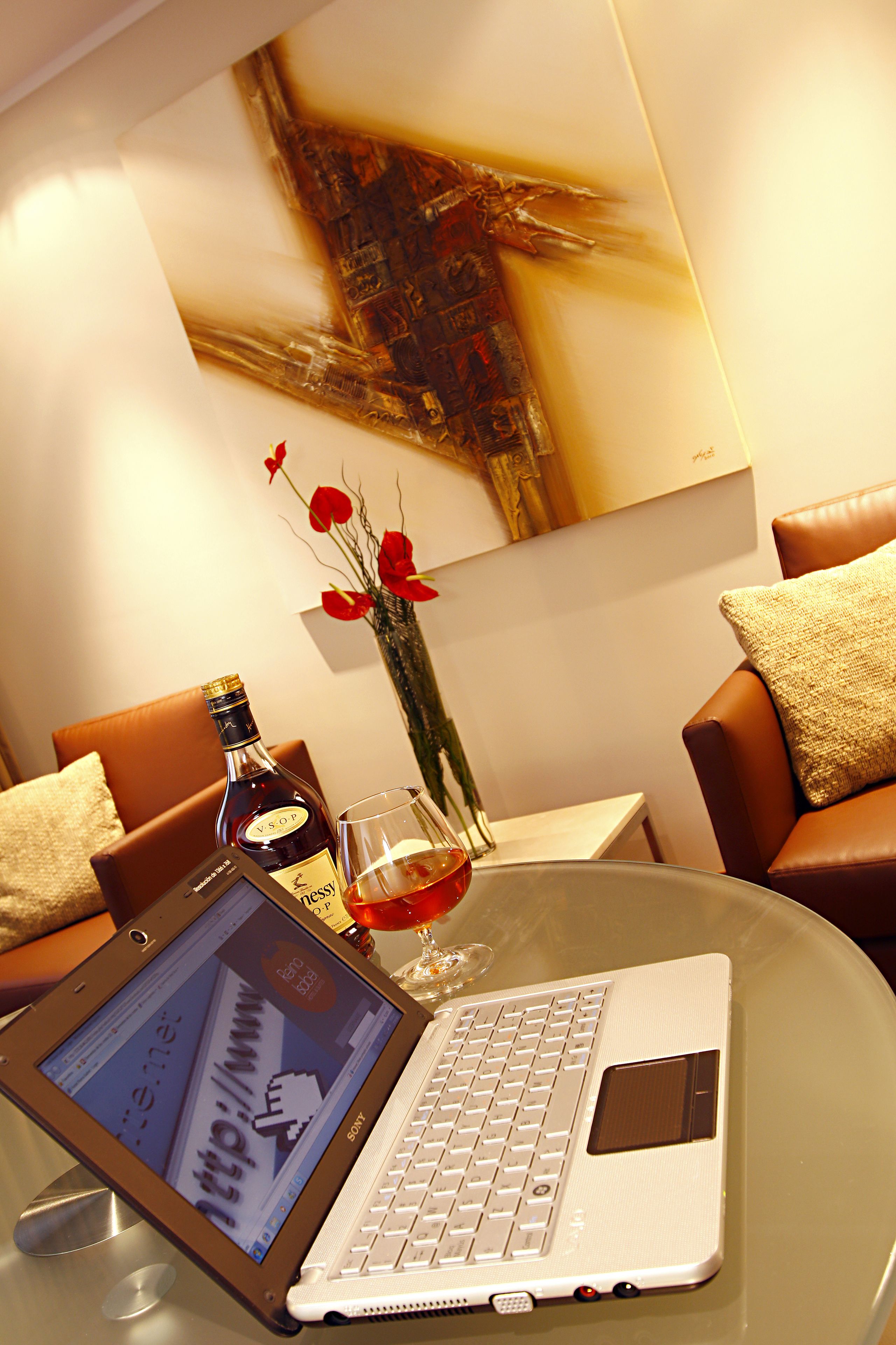 A laptop on a glass table with a bottle and glass of red wine, surrounded by brown leather chairs and a vase with red flowers.