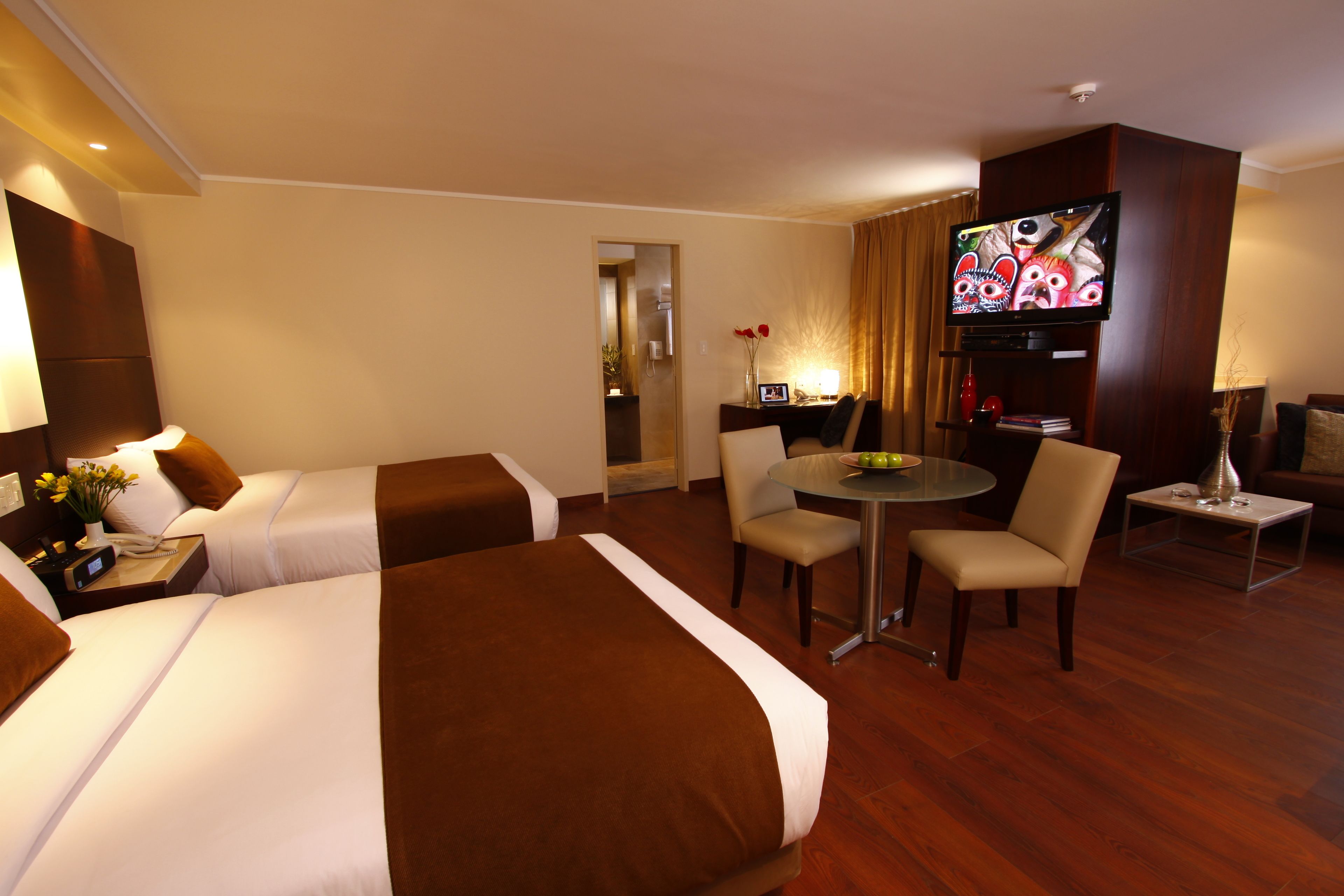 A hotel room with two beds, a television, a dining table with chairs, and a small side table with a lamp and flowers.