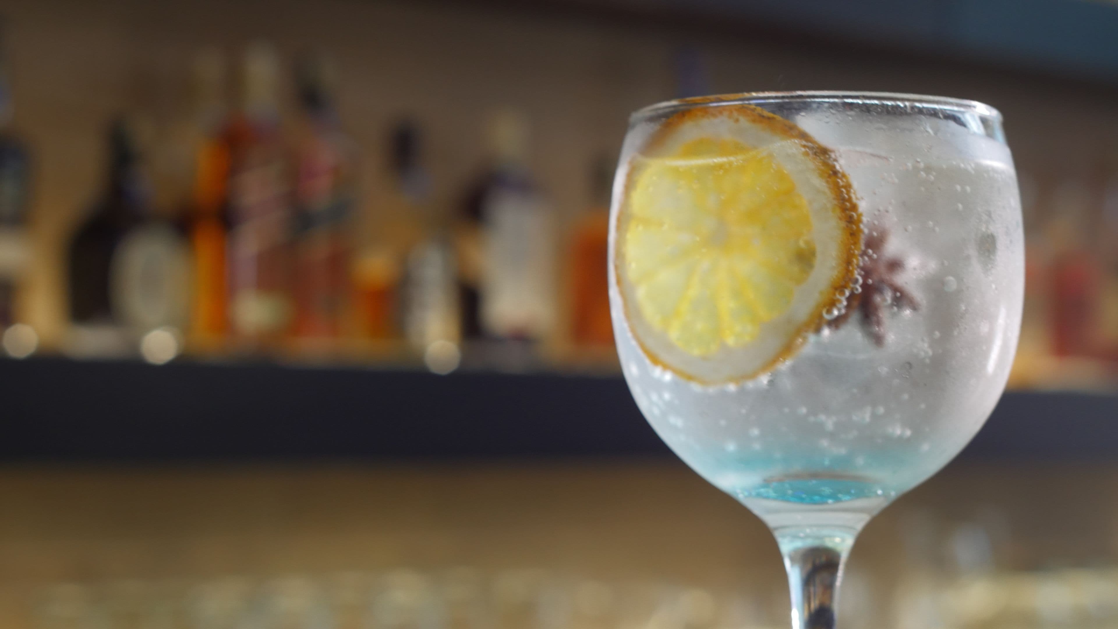 A close-up of a cocktail glass with a lemon slice garnish, ice cubes, and a blue liquid, likely a cocktail, with a blurred bar background.