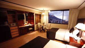 Habitación de hotel con dos camas, un escritorio con silla, una mesita de noche con lámpara y vistas a un paisaje urbano con montaña.