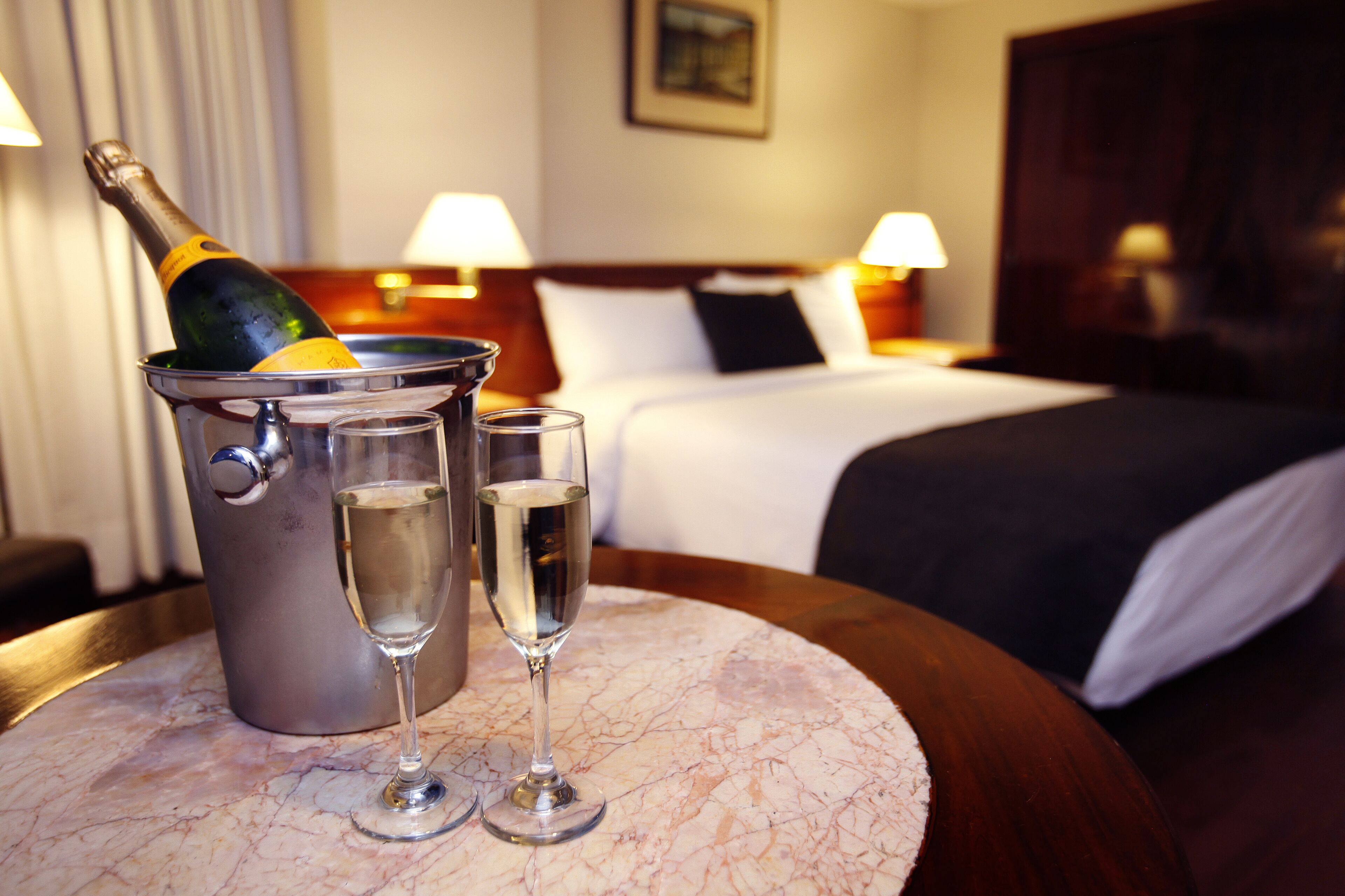A hotel room with a bed, bedside lamps, a marble table with a champagne bottle and two glasses.