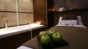 Una habitación de hotel moderna con una cama grande, una mesita de noche con una bandeja de manzanas verdes, un baño con bañera y un estante en la pared con toallas.