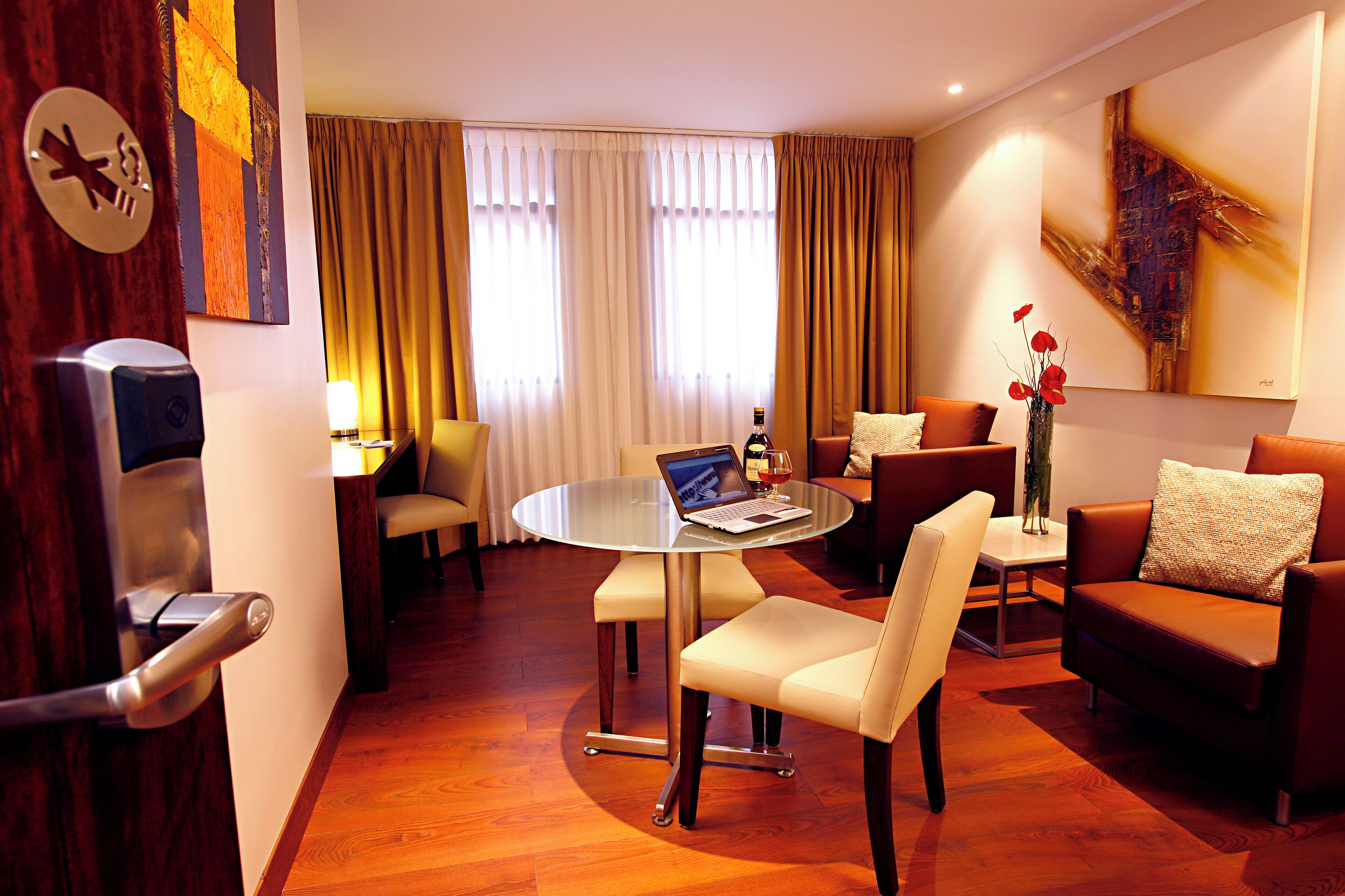 A modern hotel room with a round glass table, leather sofas, a laptop on the table, and a large painting on the wall.