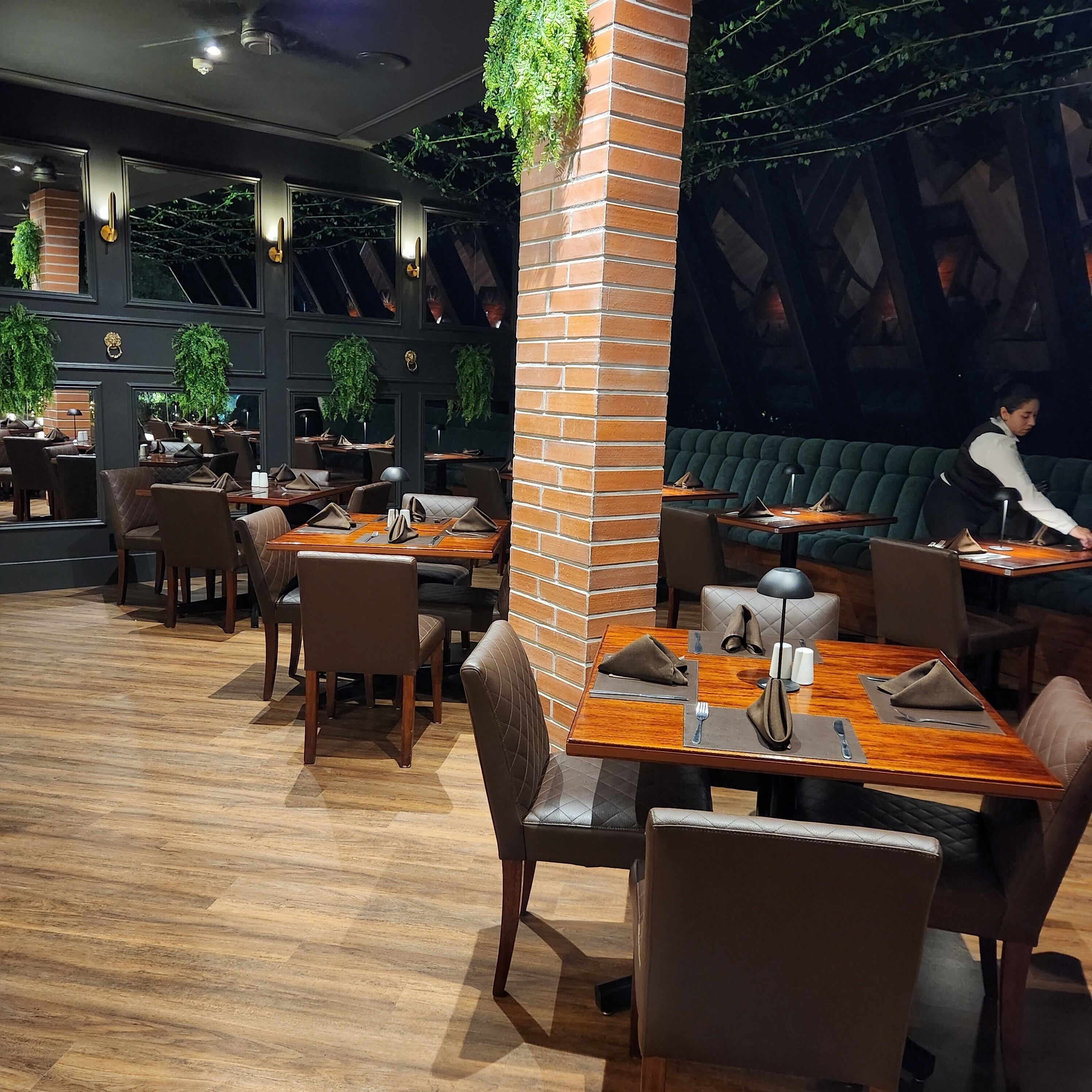 A restaurant interior with brick pillars, wooden tables, leather chairs, and hanging plants.