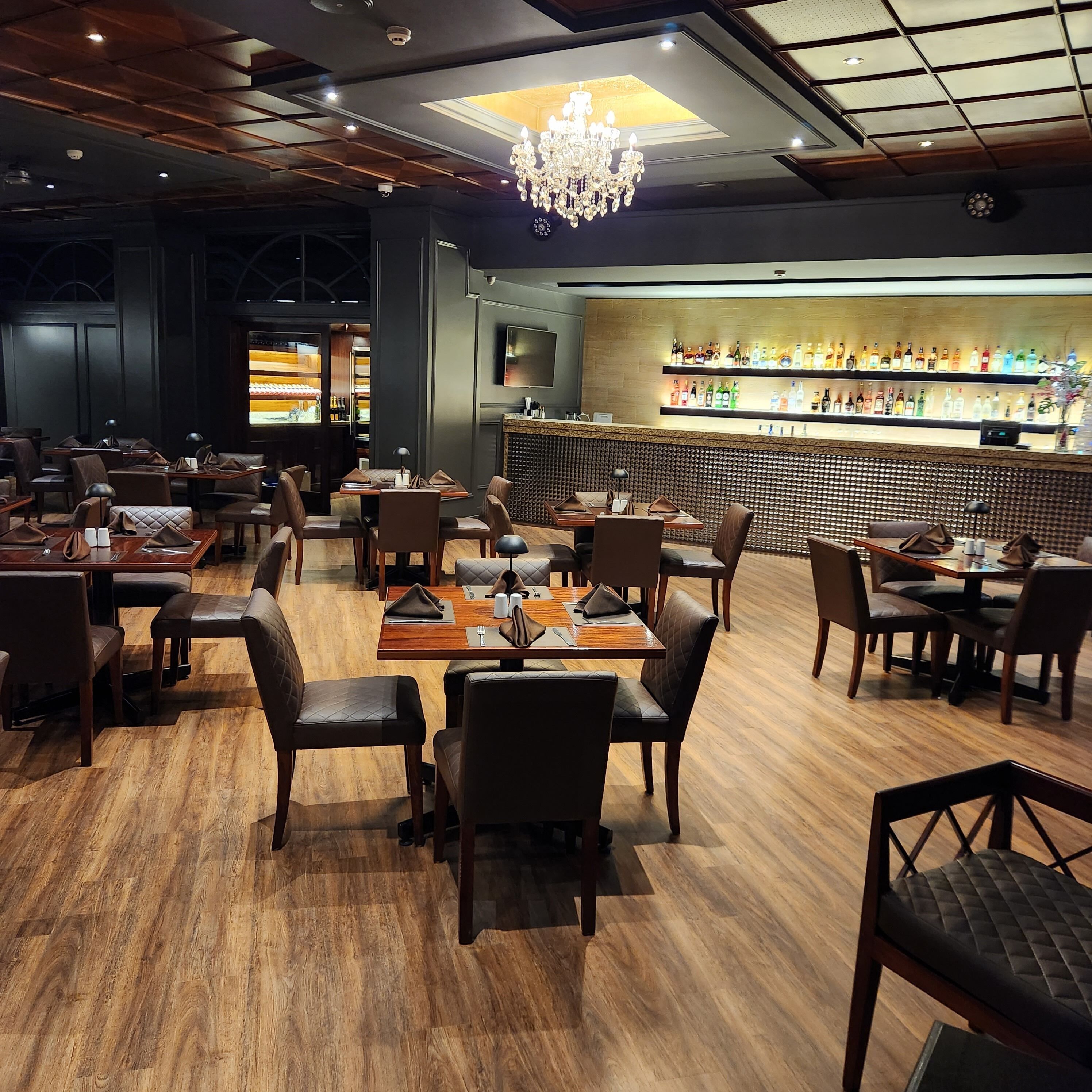 A restaurant with wooden flooring, tables with chairs, a bar area with shelves of bottles, and a chandelier.
