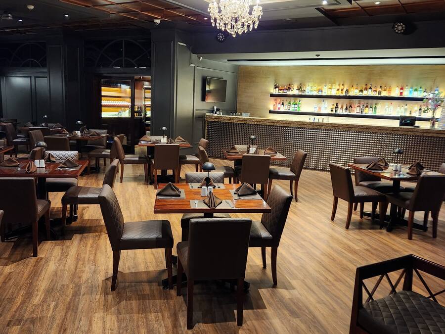 A restaurant with wooden flooring, tables with chairs, a bar area with shelves of bottles, and a chandelier.