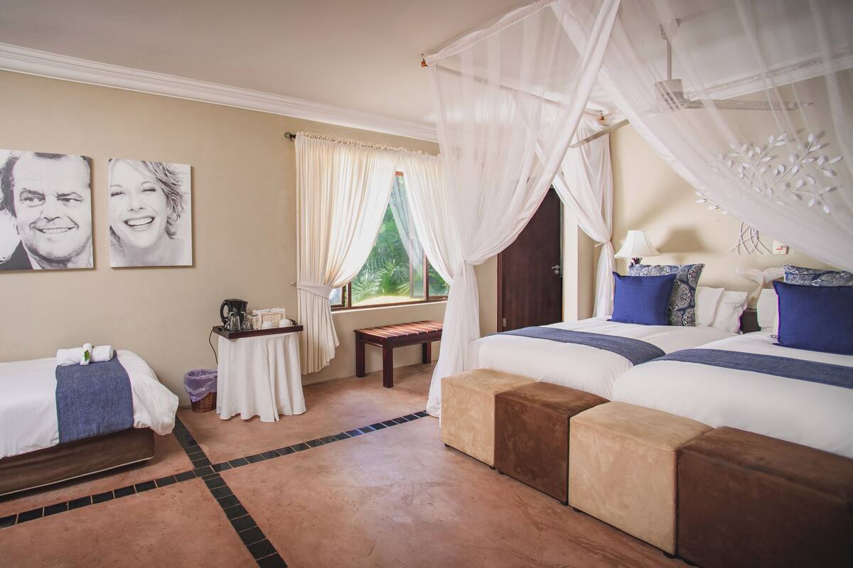 A bedroom with two beds, a bench, a small table with a coffee maker, and a window with curtains.