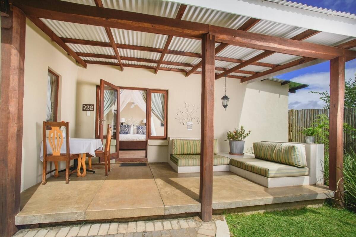 A covered outdoor area with a wooden pergola, a dining table with chairs, a sofa, and a bed with a canopy.