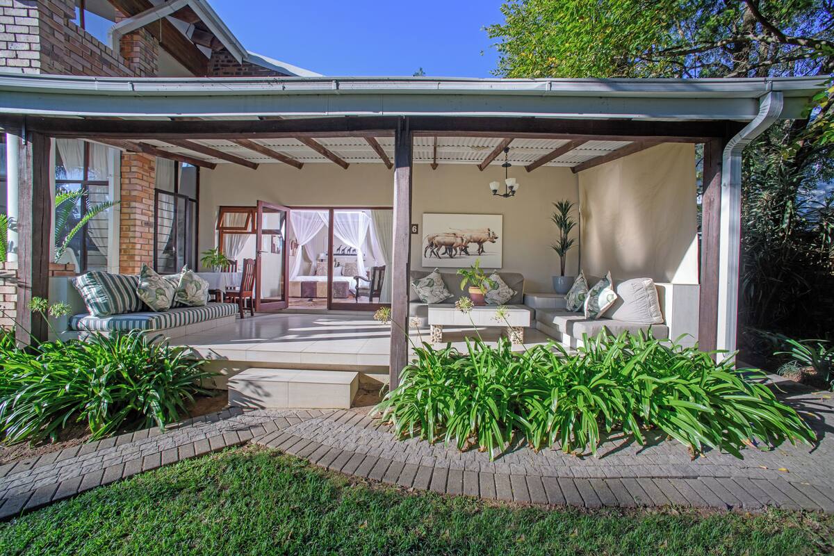 A covered outdoor seating area with a sofa and chairs, surrounded by potted plants and a brick pathway.
