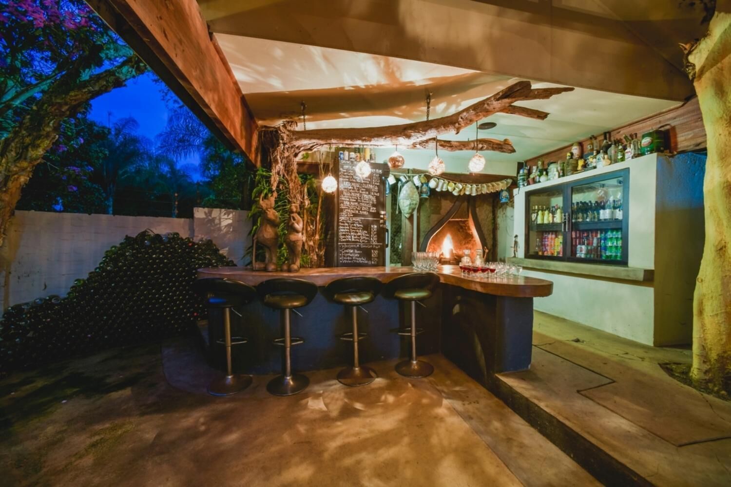 An outdoor bar area with a rustic design, including a wooden bar counter, three stools, hanging lights, and a brick wall with a chalkboard menu.