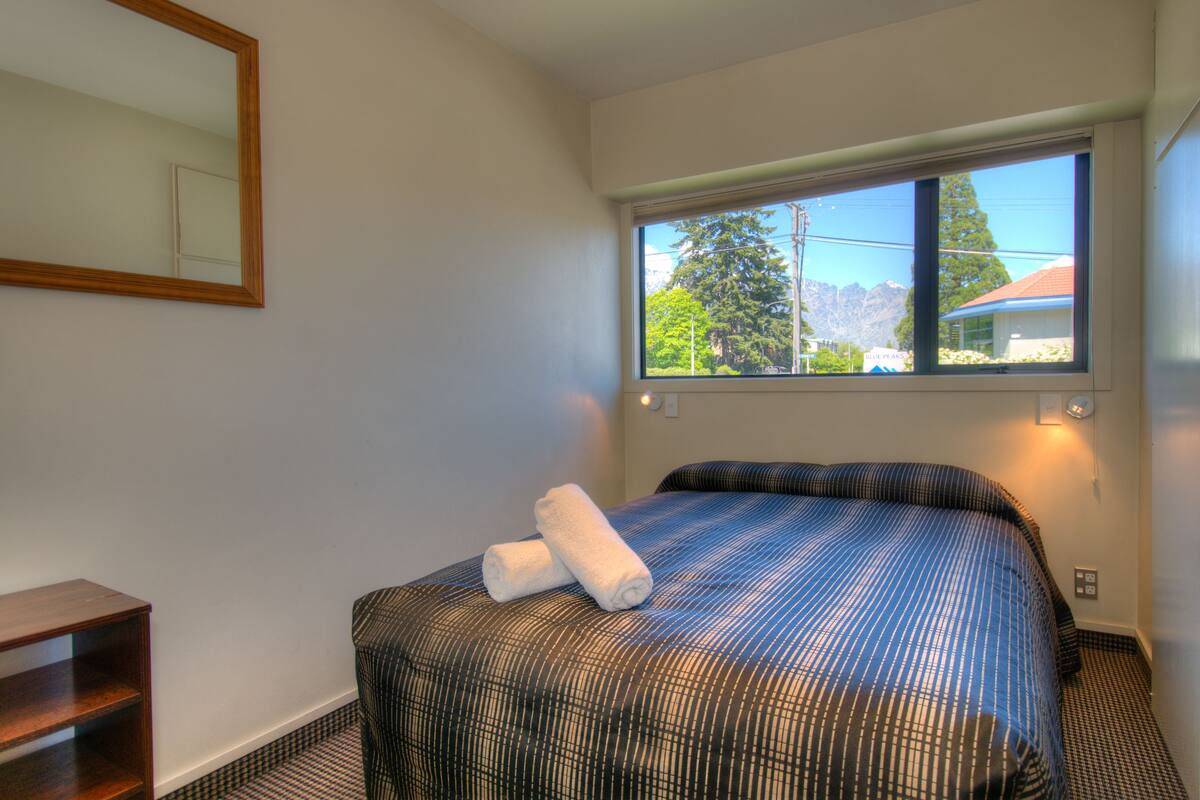 A hotel room with a bed, a window with a mountain view, a wooden nightstand, and a mirror on the wall.