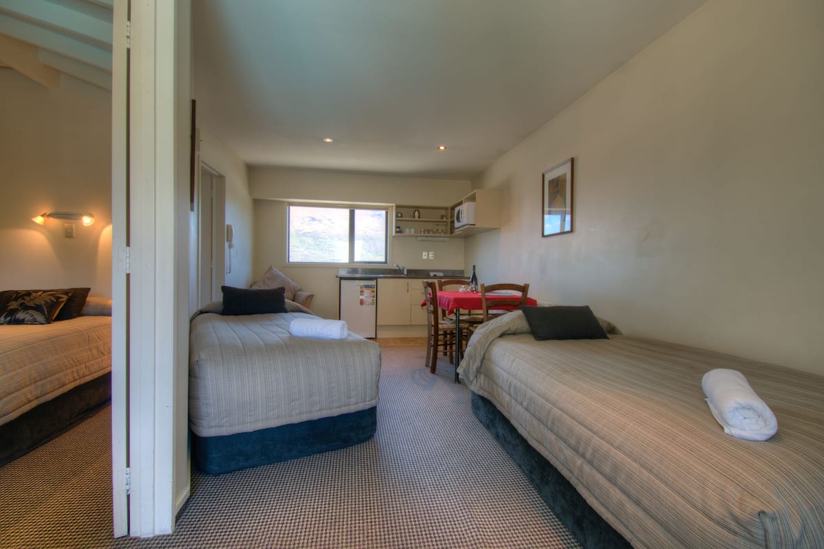 A compact hotel room with two beds, a small kitchenette, and a dining area.