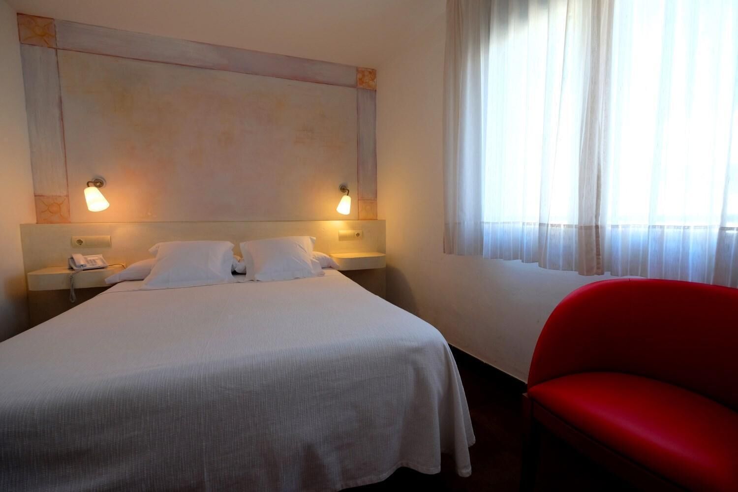Ein Hotelzimmer mit einem großen Bett, zwei Nachttischlampen, einem Telefon und einem roten Sessel.