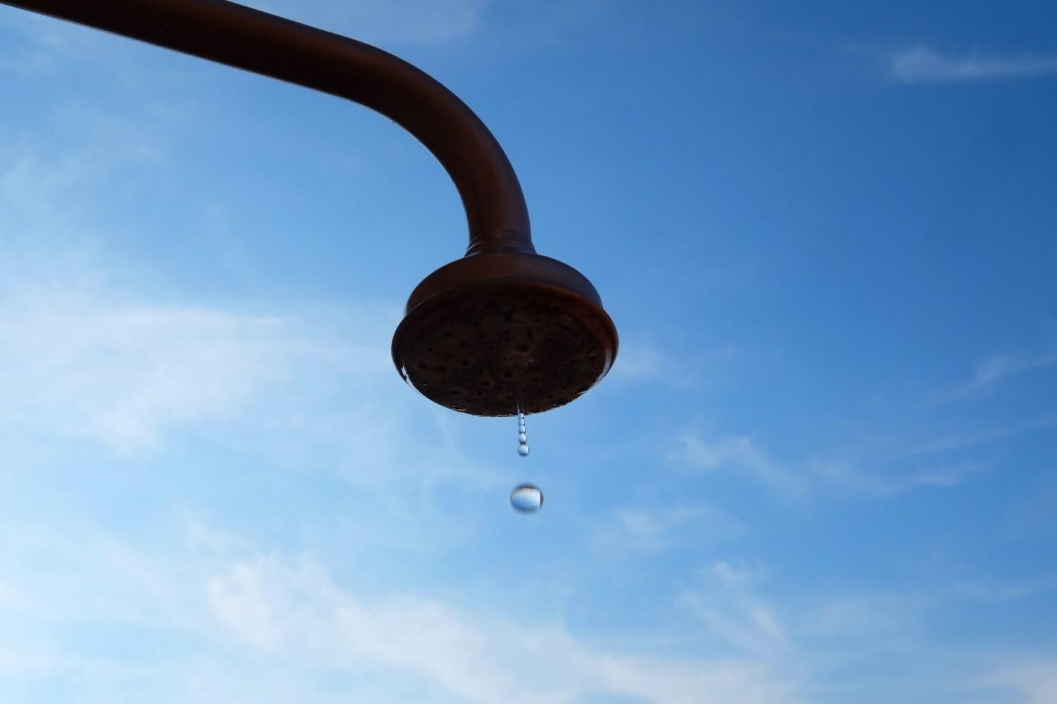 Un pommeau de douche dont l’eau s’écoule goutte à goutte, sur fond de ciel bleu clair.