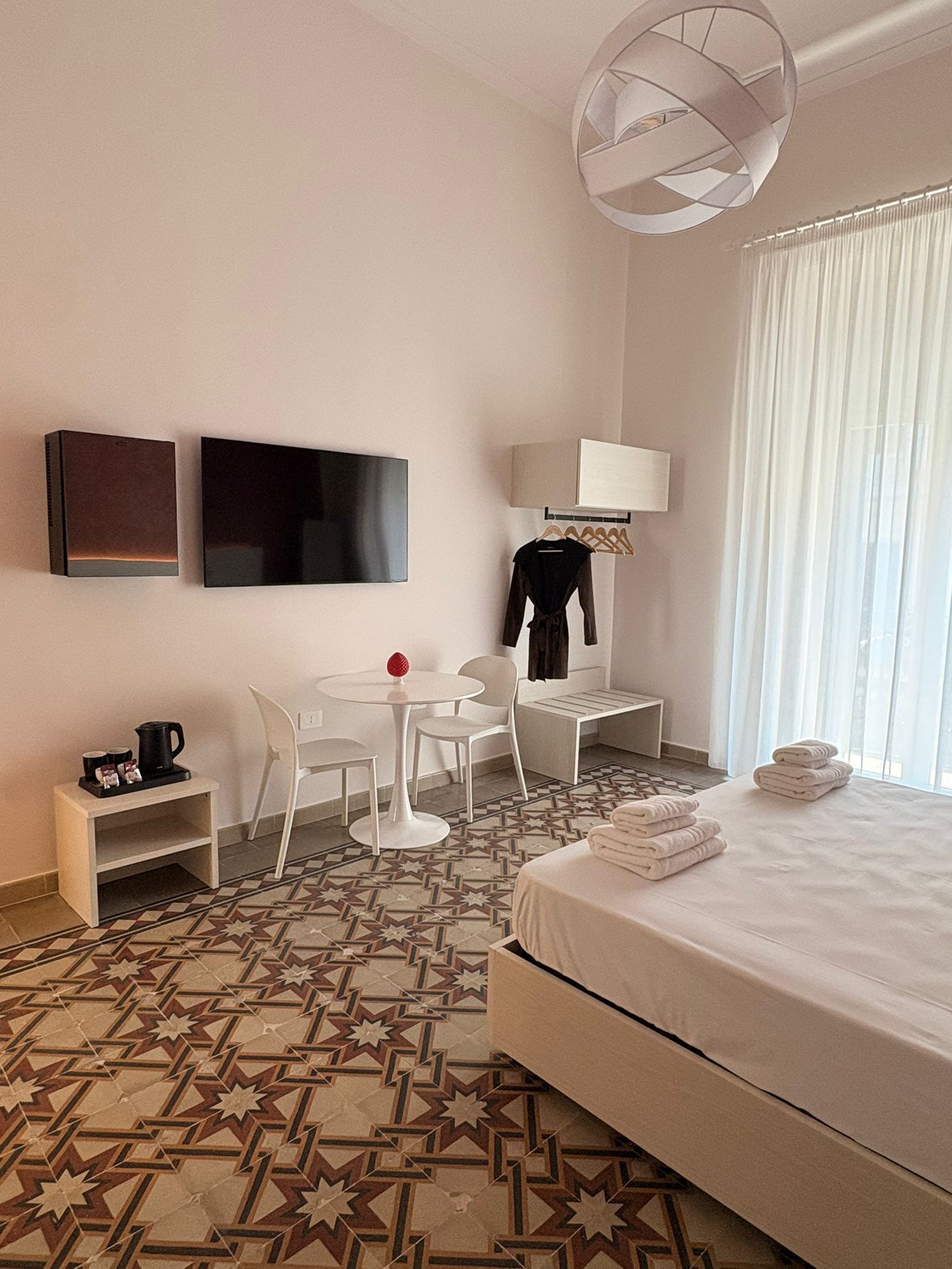 Habitación de hotel con cama, escritorio, silla, mesita, lámpara, televisor y ventana con cortinas.