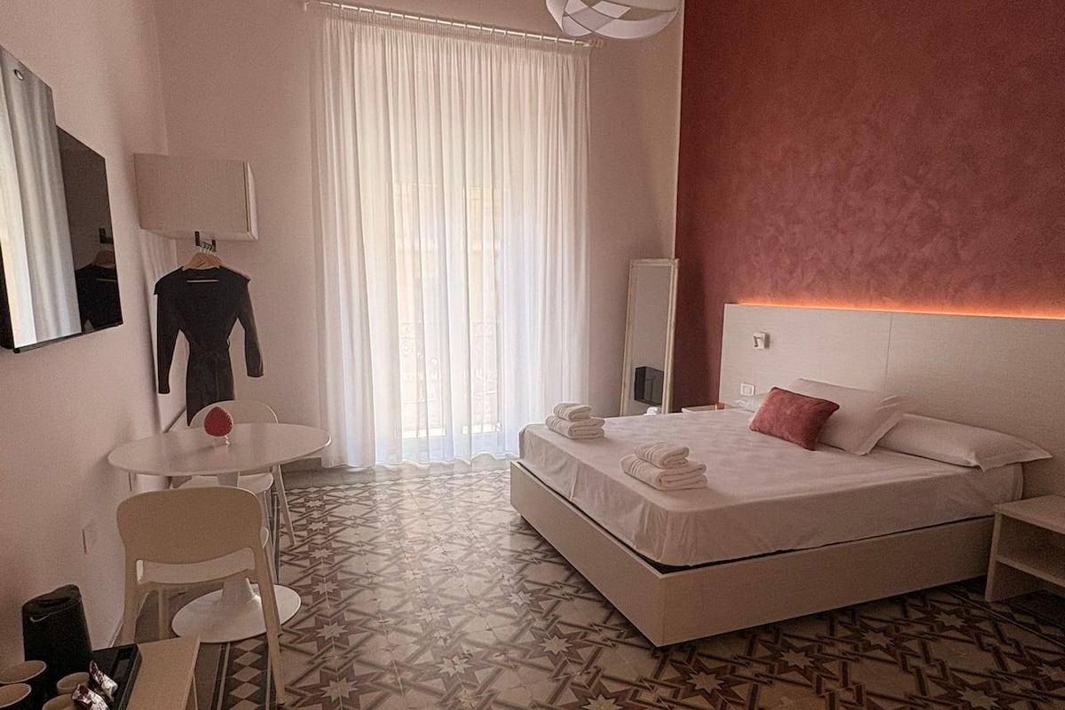 A hotel room with a bed, a desk, a chair, a mirror, and a patterned floor.