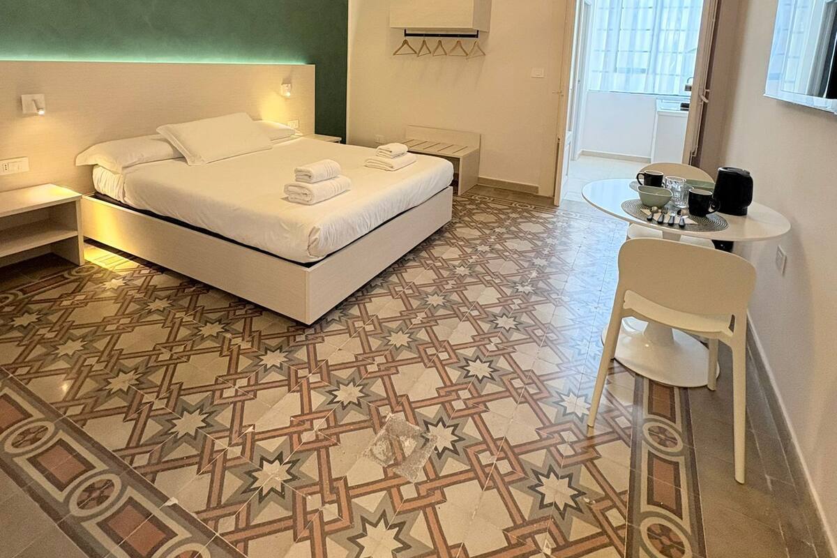 A modern bedroom with a bed, bedside tables, a hanging light fixture, a small table with cups and a kettle, and a patterned floor.