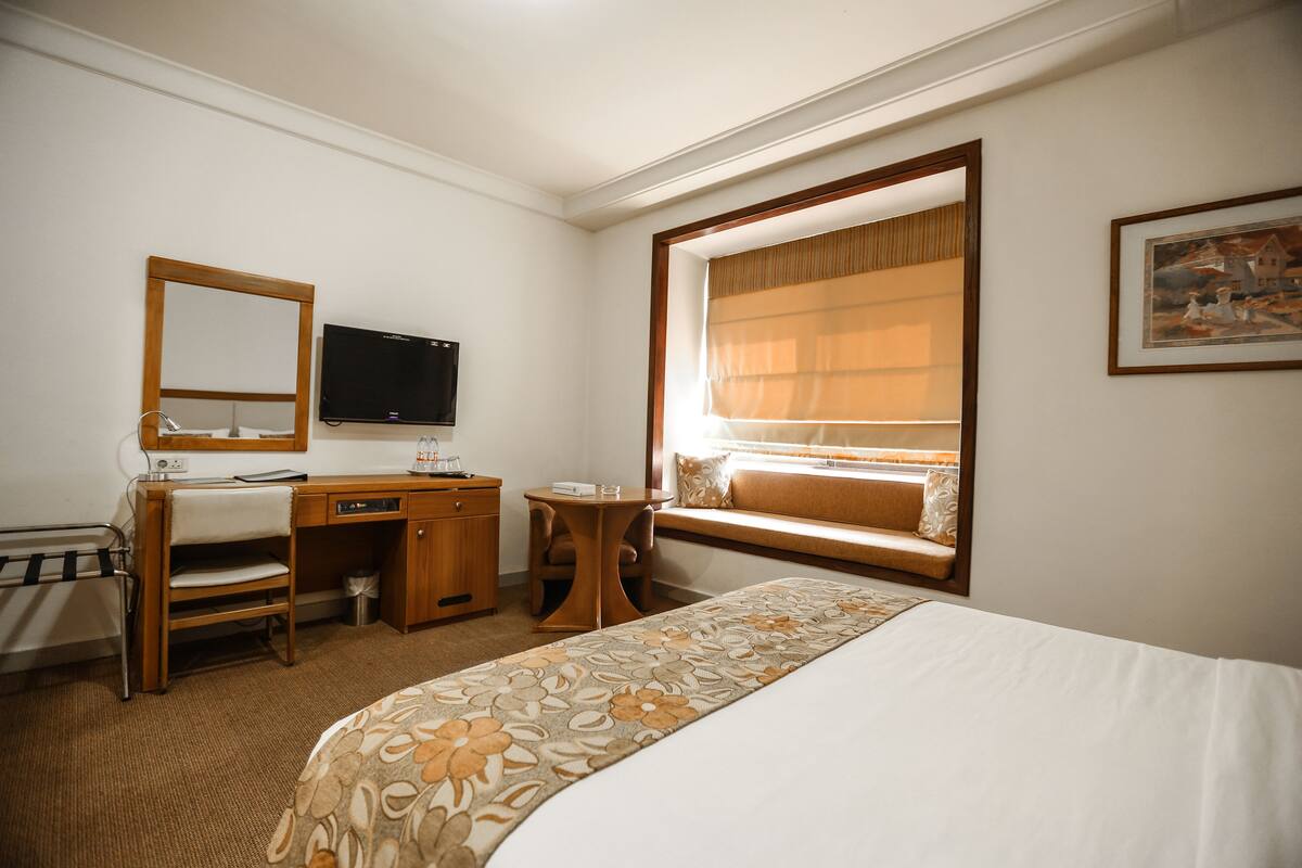 A hotel room with a bed, a desk, a chair, a television, a window with a view, and a painting on the wall.