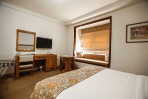 A hotel room with a bed, a desk, a chair, a television, a window with a view, and a painting on the wall.