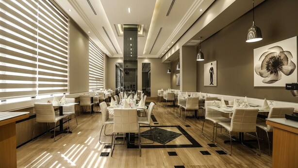 A modern hotel dining area with multiple tables and chairs, a central column, and artwork on the walls. - Gardenia Hotel (Amman)