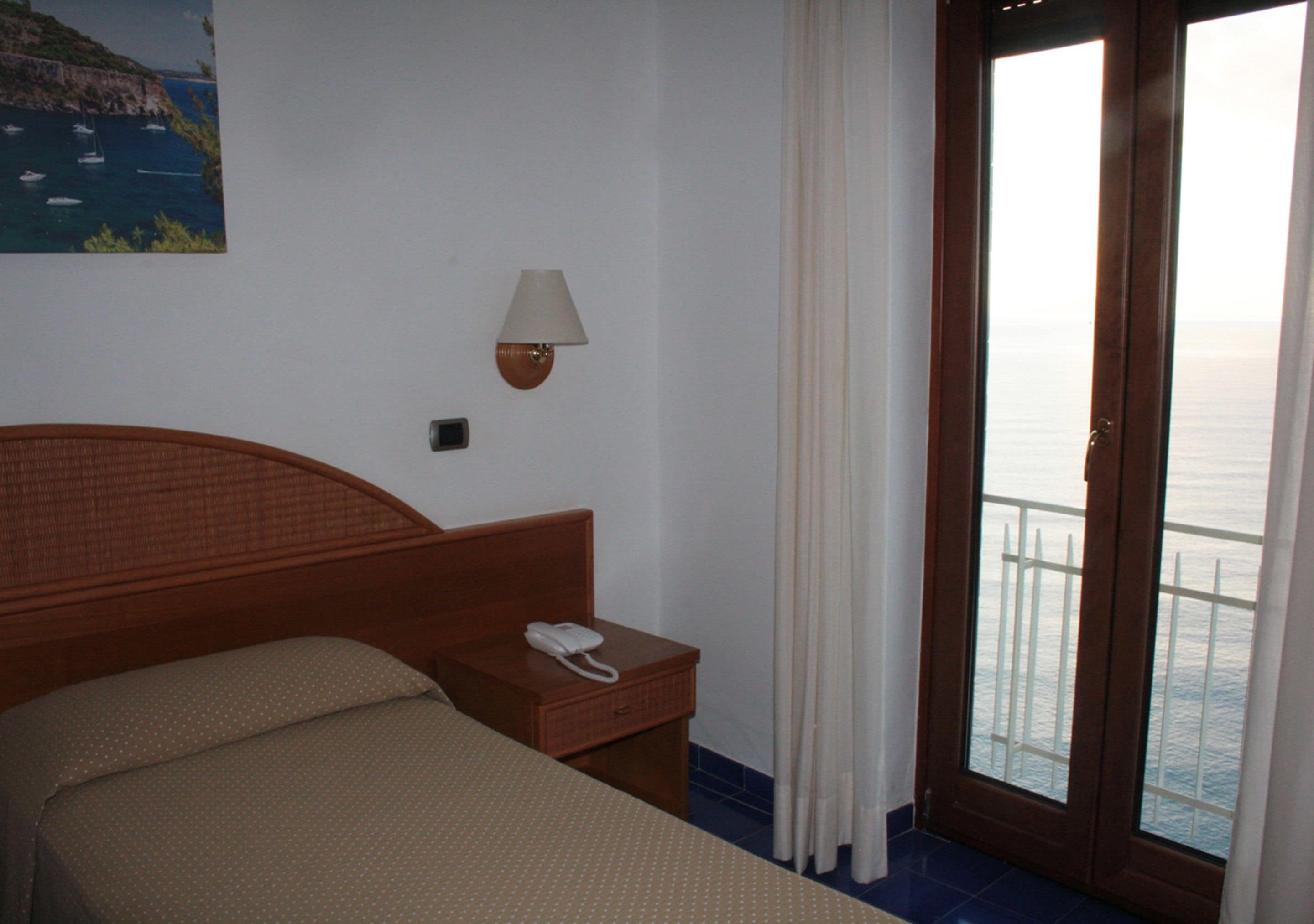 A hotel room with a bed, a bedside table with a phone, a wall-mounted lamp, and a view of the sea through a door.