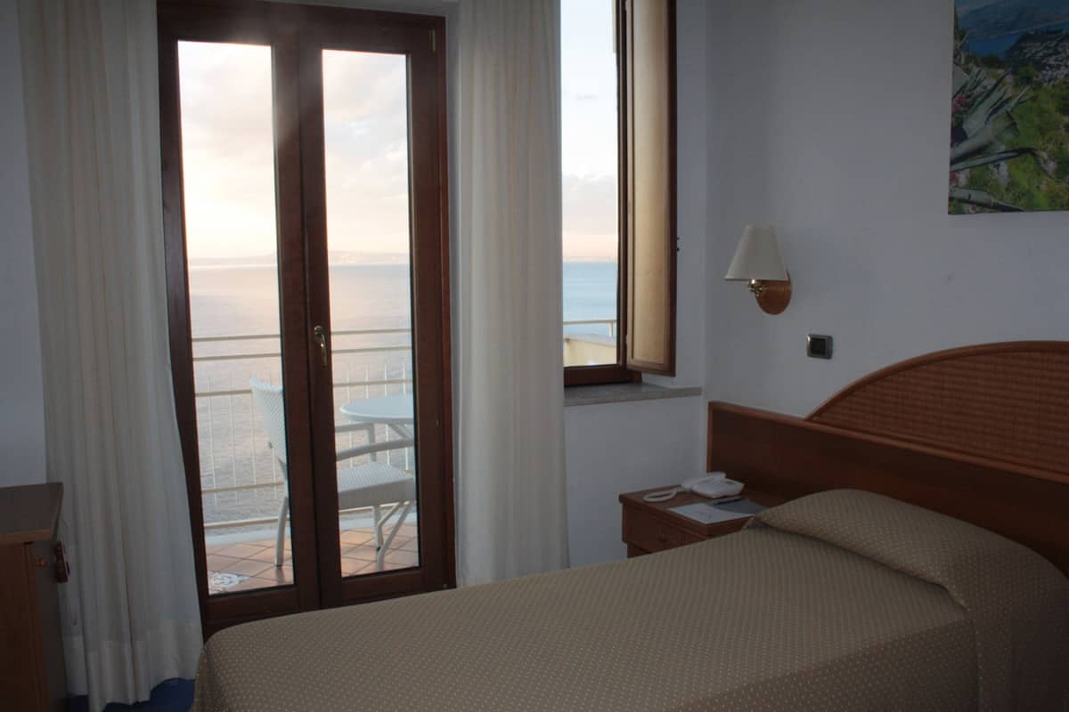 Ein Hotelzimmer mit Bett, Nachttisch, Lampe, einem Gemälde und Meerblick durch ein Fenster.