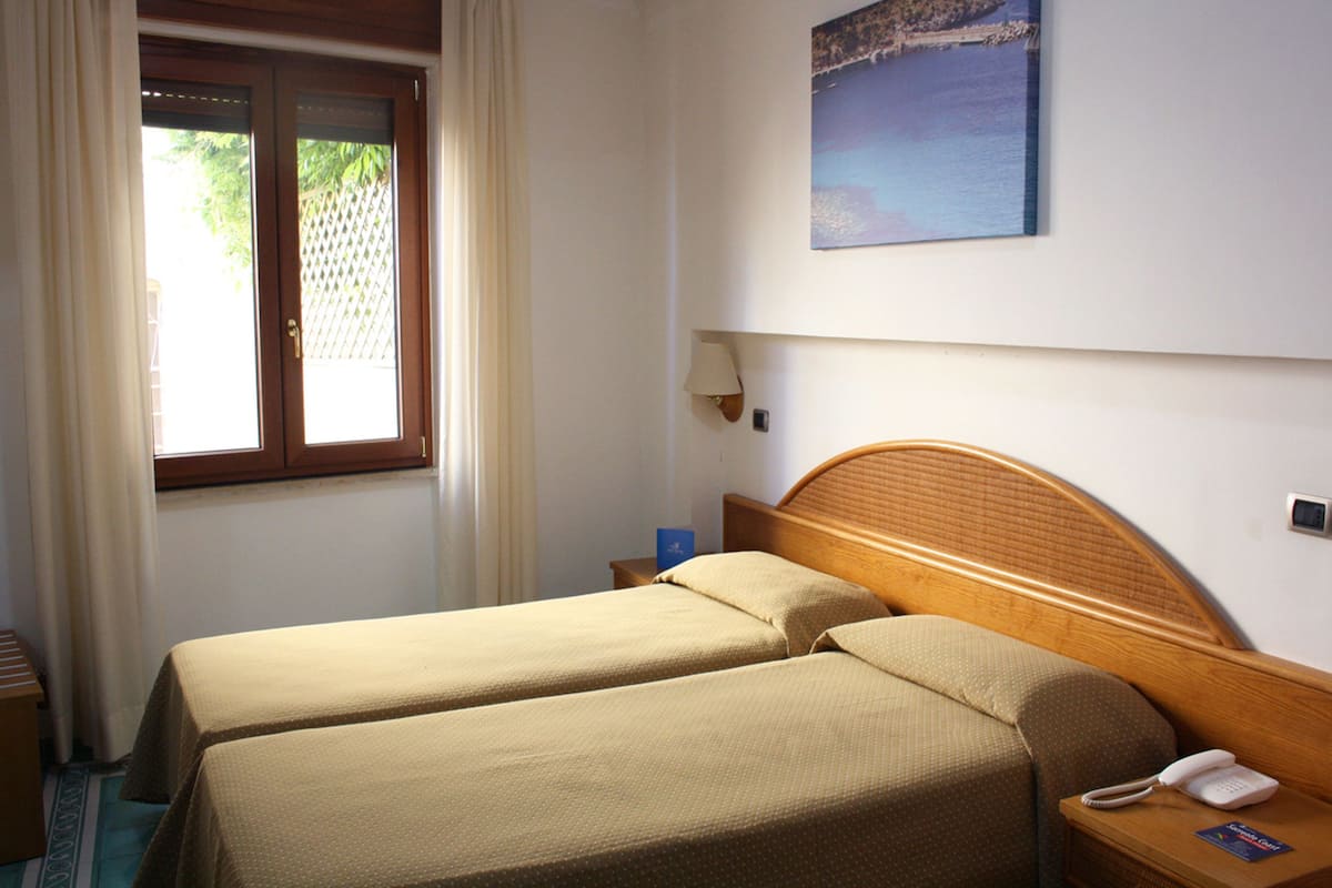 Ein Hotelzimmer mit zwei Betten, einem Fenster mit Vorhängen, einem Nachttisch mit Lampe und einem Bild an der Wand.